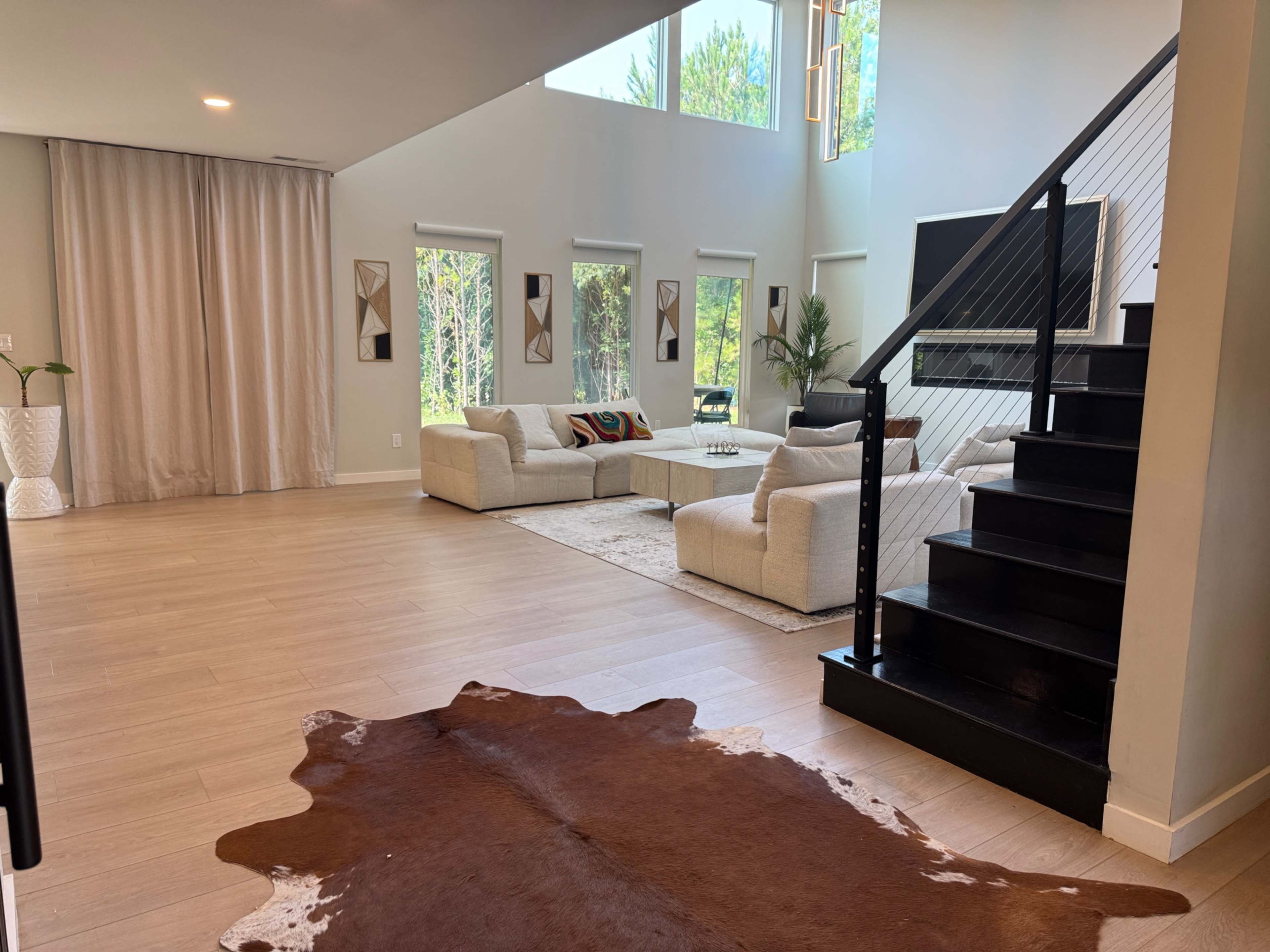 A modern living room features a large sectional sofa, a rug, and large windows allowing natural light, with a staircase in the background.