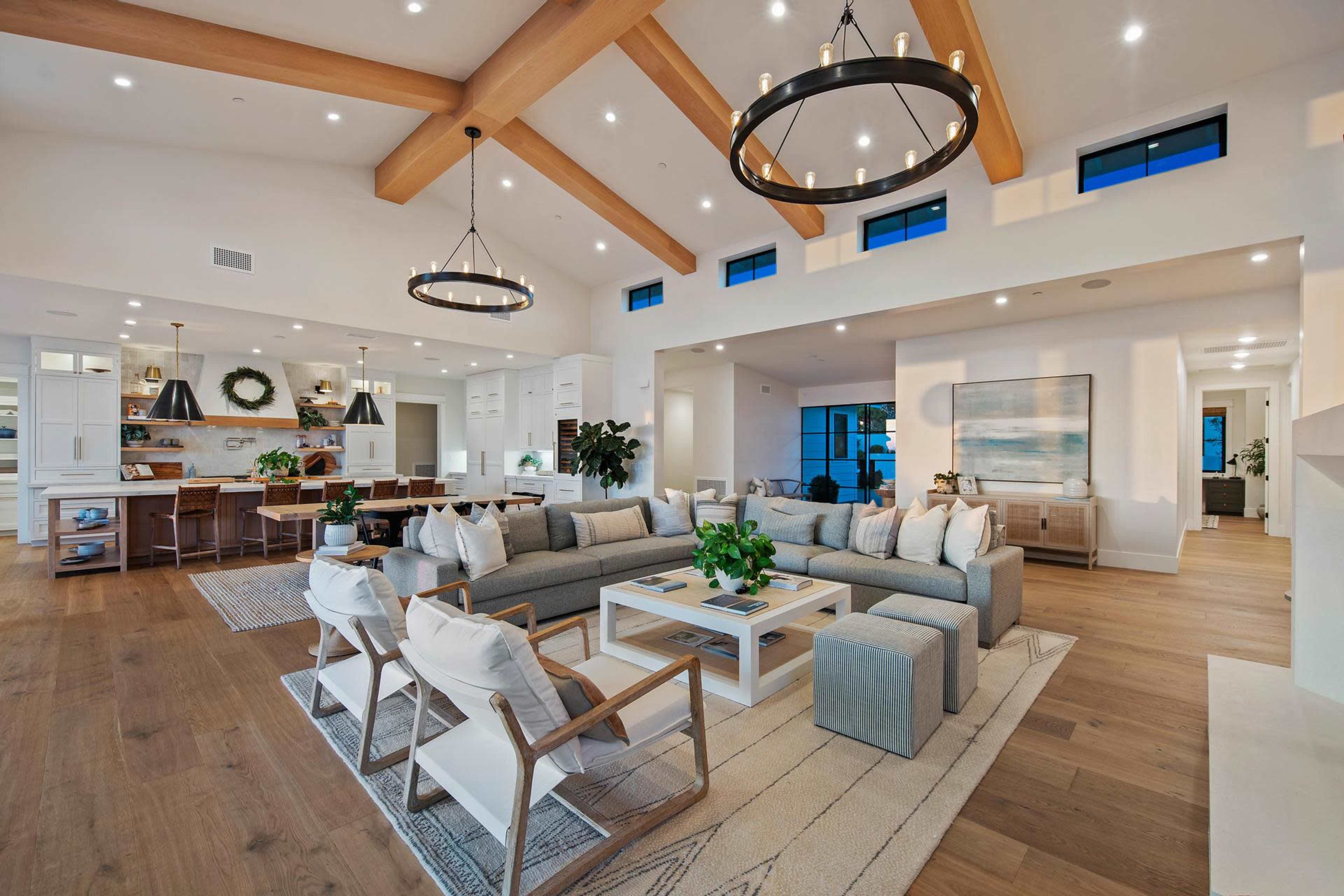 The image shows a spacious living area with a light color palette, featuring a large sectional sofa, wooden beams on the ceiling, and a dining area in the background.