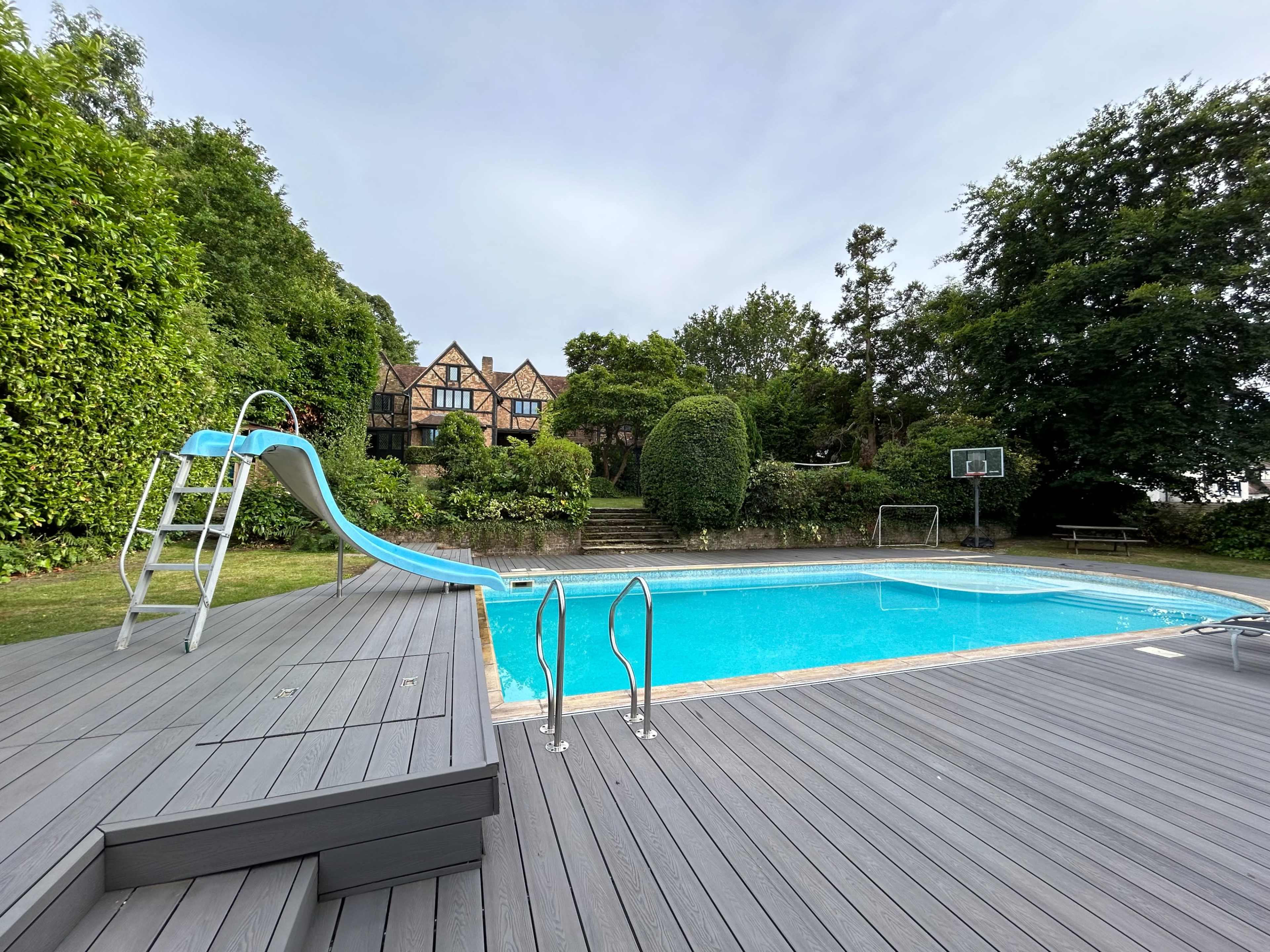 The image shows a swimming pool with a slide, surrounded by greenery and a house in the background.
