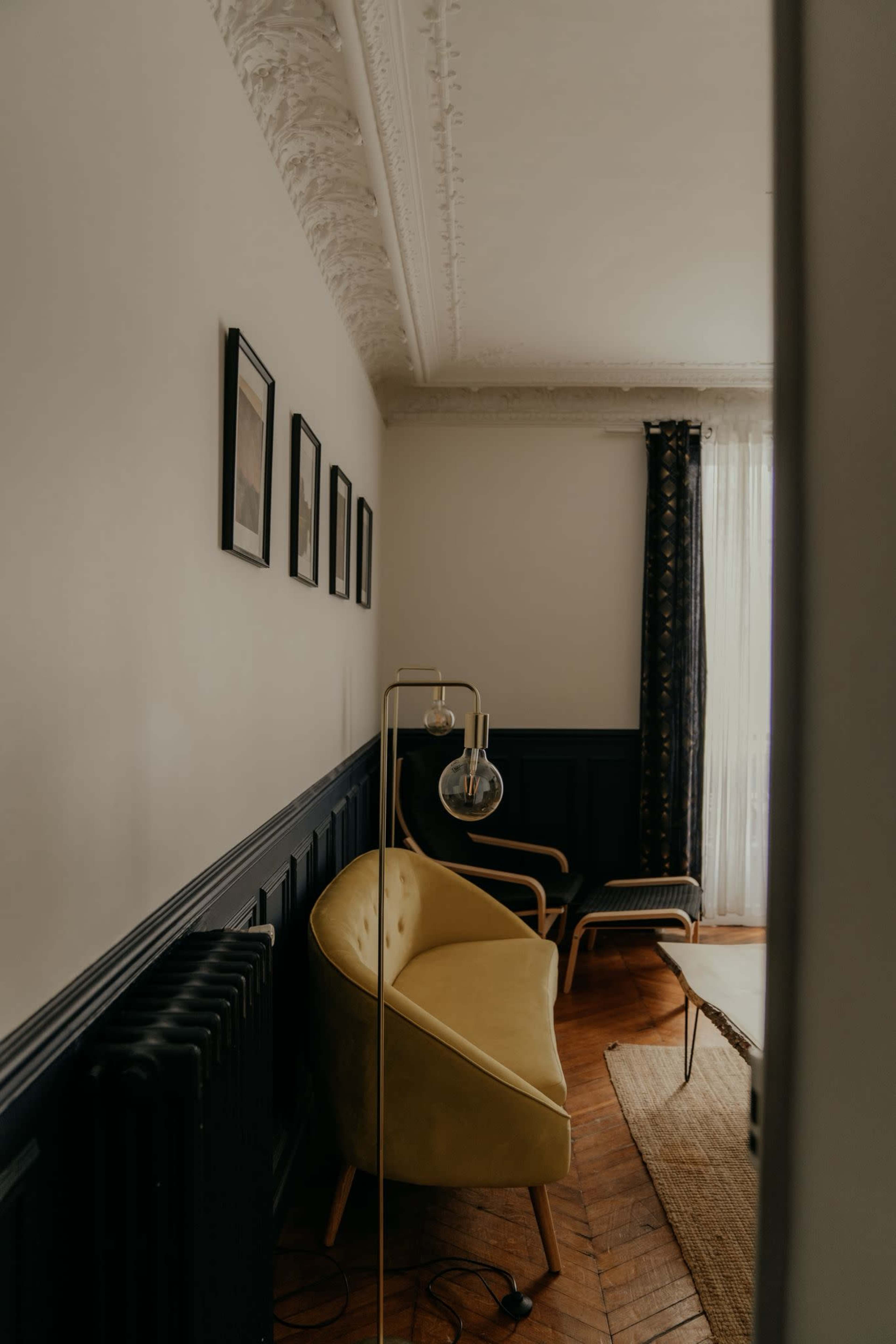 A yellow curved sofa is positioned against a dark-paneled wall, complemented by framed pictures, a floor lamp, and a view of a window with drapes.