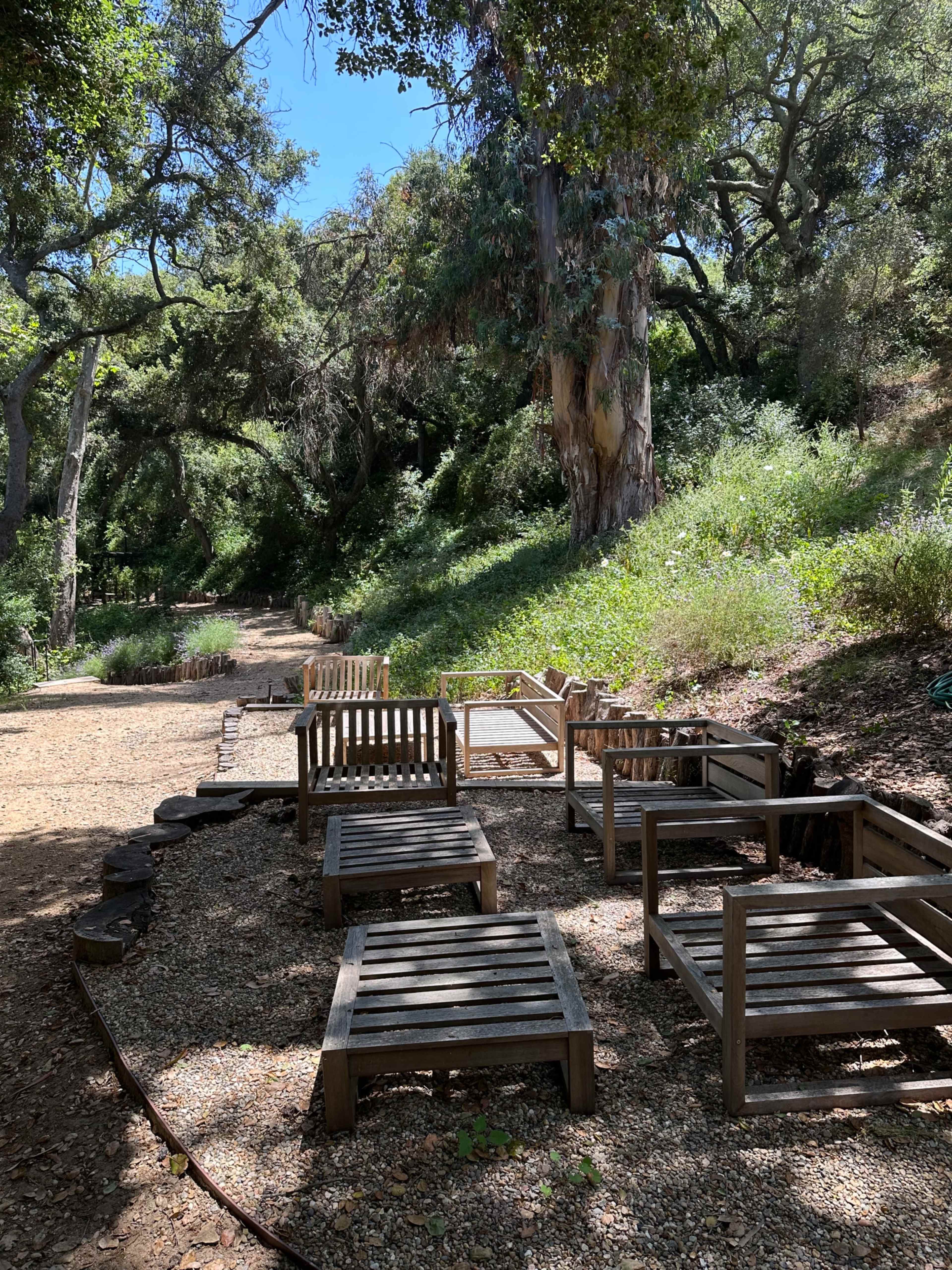 The image shows a series of wooden lounge chairs arranged on a gravel path surrounded by greenery and trees.