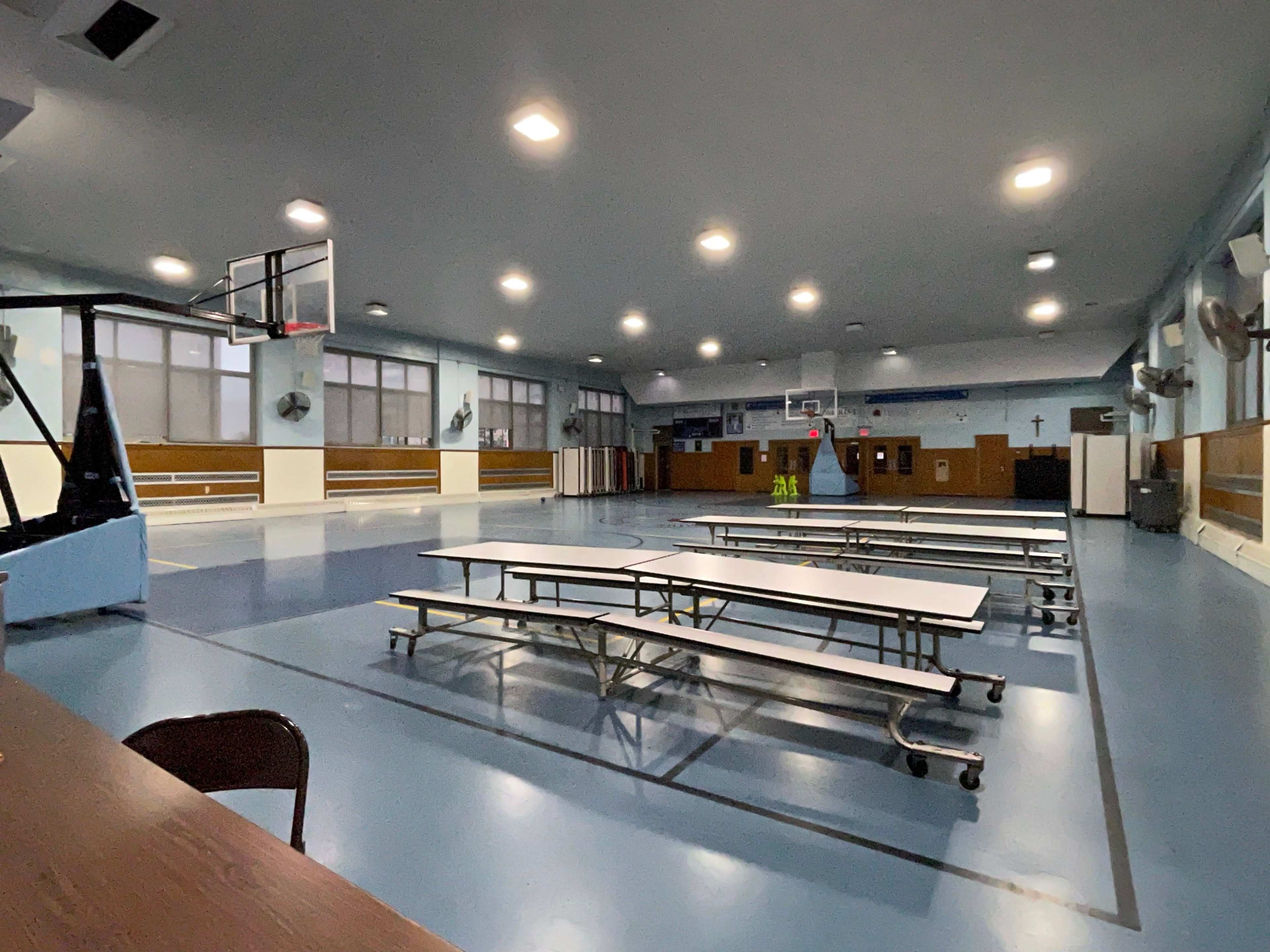 An empty gymnasium with basketball hoops, several long tables arranged in rows, and bright overhead lights.