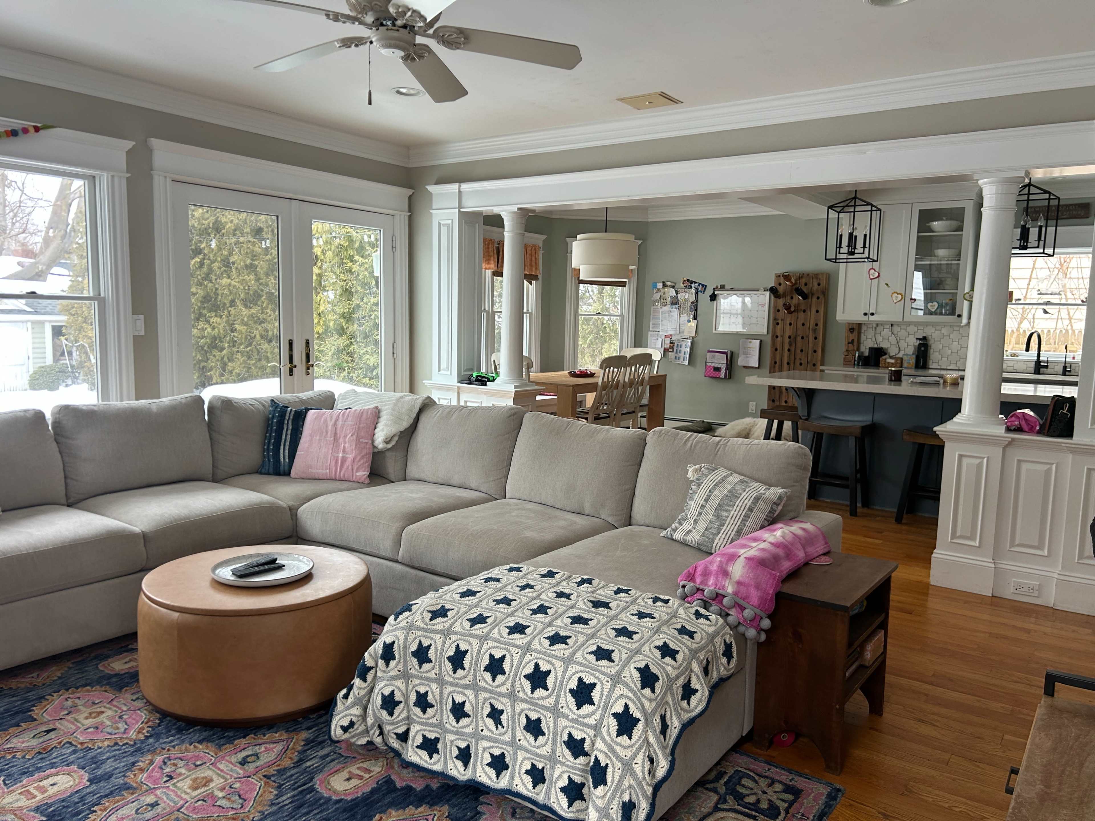 The image shows a spacious living room with a large sectional couch, a round coffee table, and a view into a connected kitchen area featuring a dining table and bar stools.