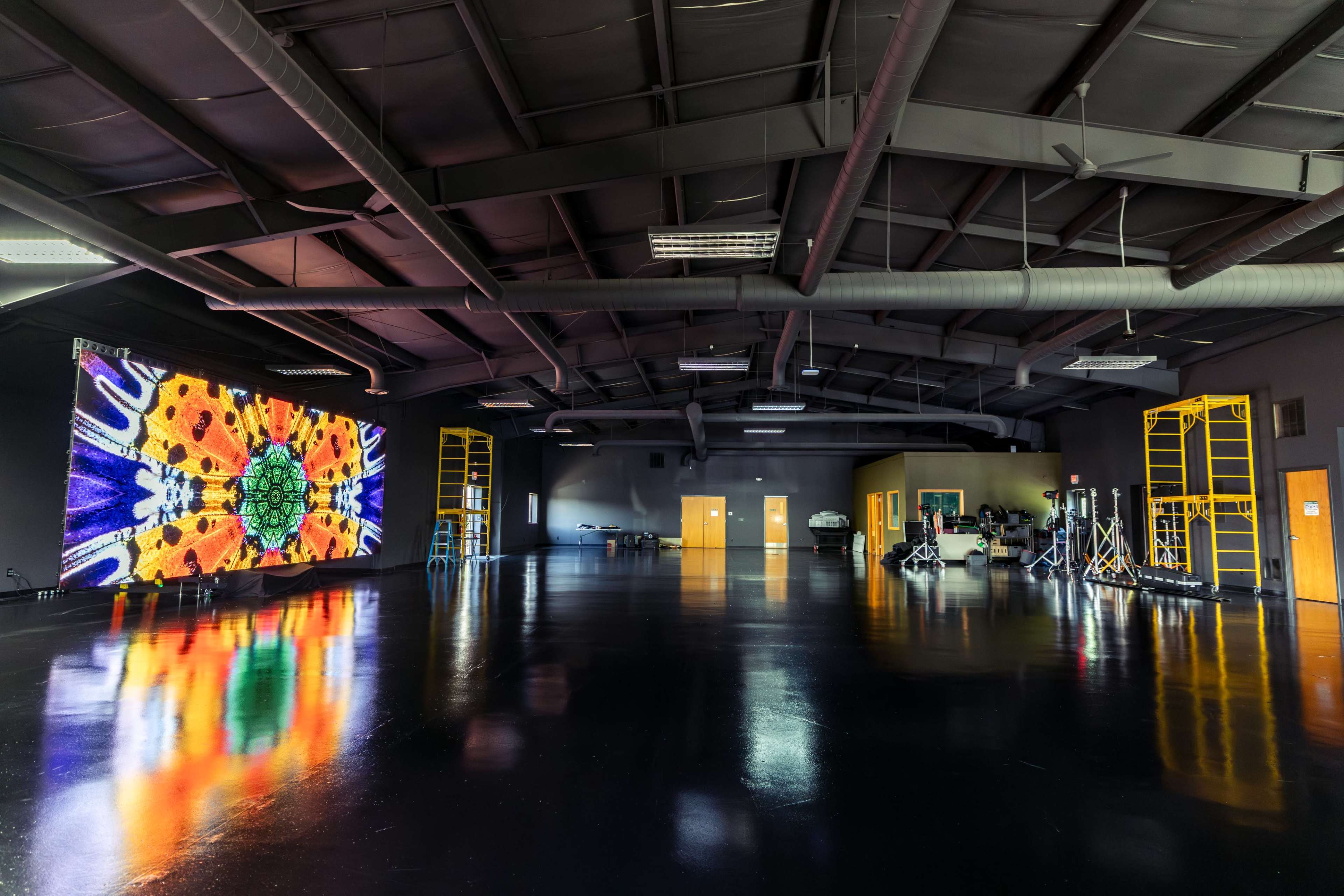 The image shows a spacious industrial room with shiny black floors, featuring a large colorful digital display on one wall and various equipment set up for audiovisual production.