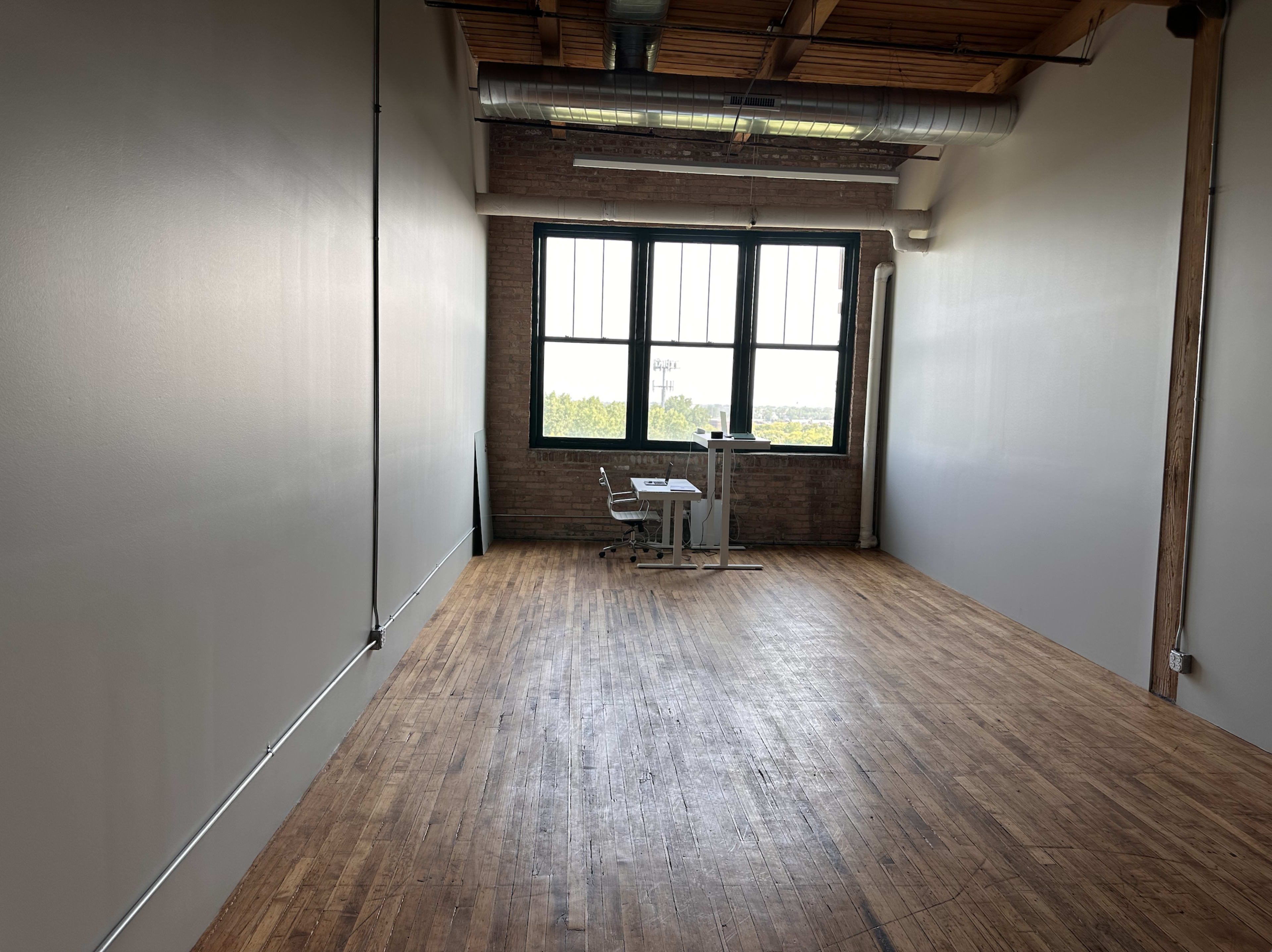 A spacious, empty room with wooden flooring, large windows, and a small desk with a chair against one wall.