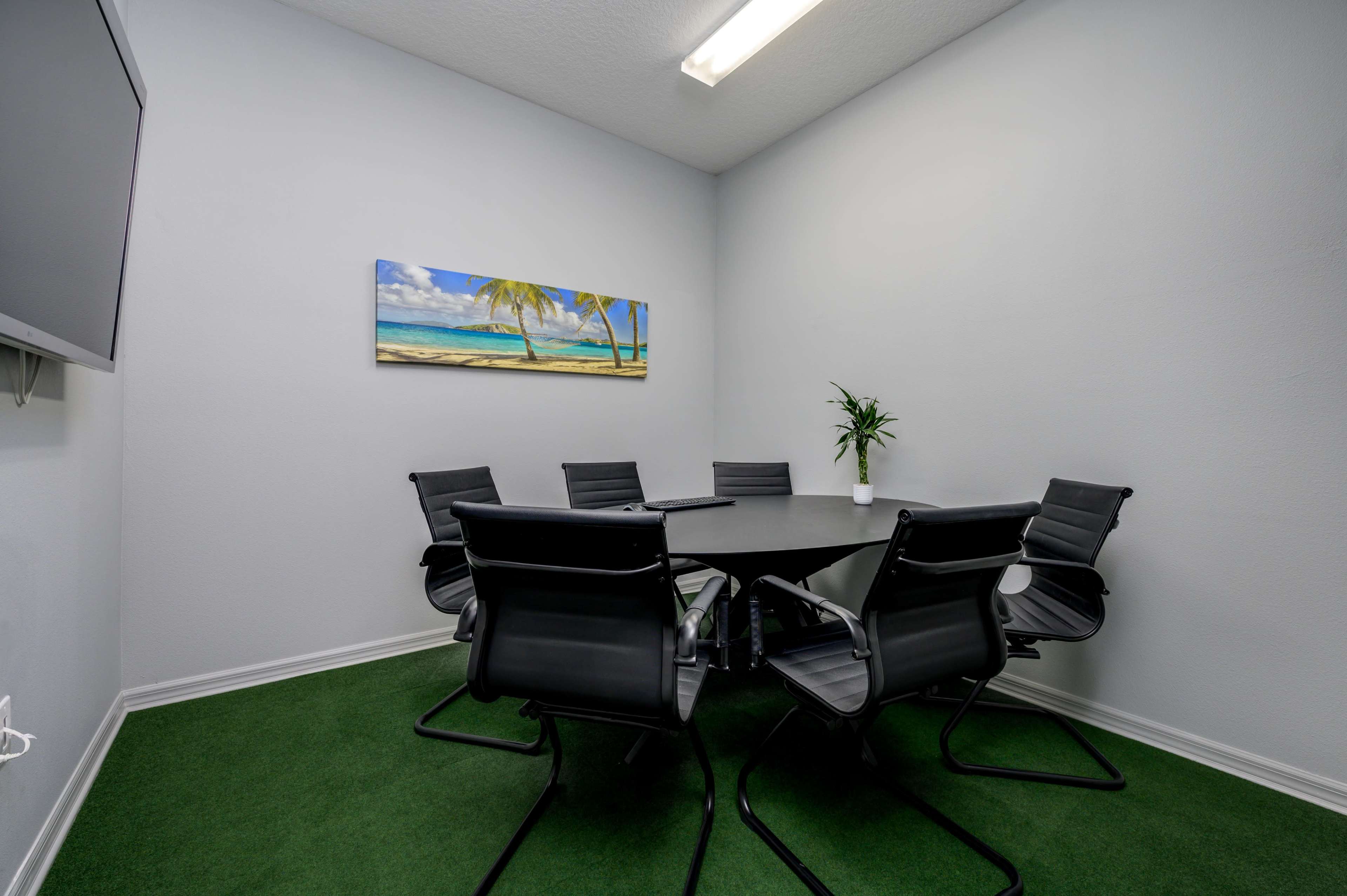A small conference room features a round table surrounded by six black chairs, with a tropical landscape painting on the wall and a plant in the corner.