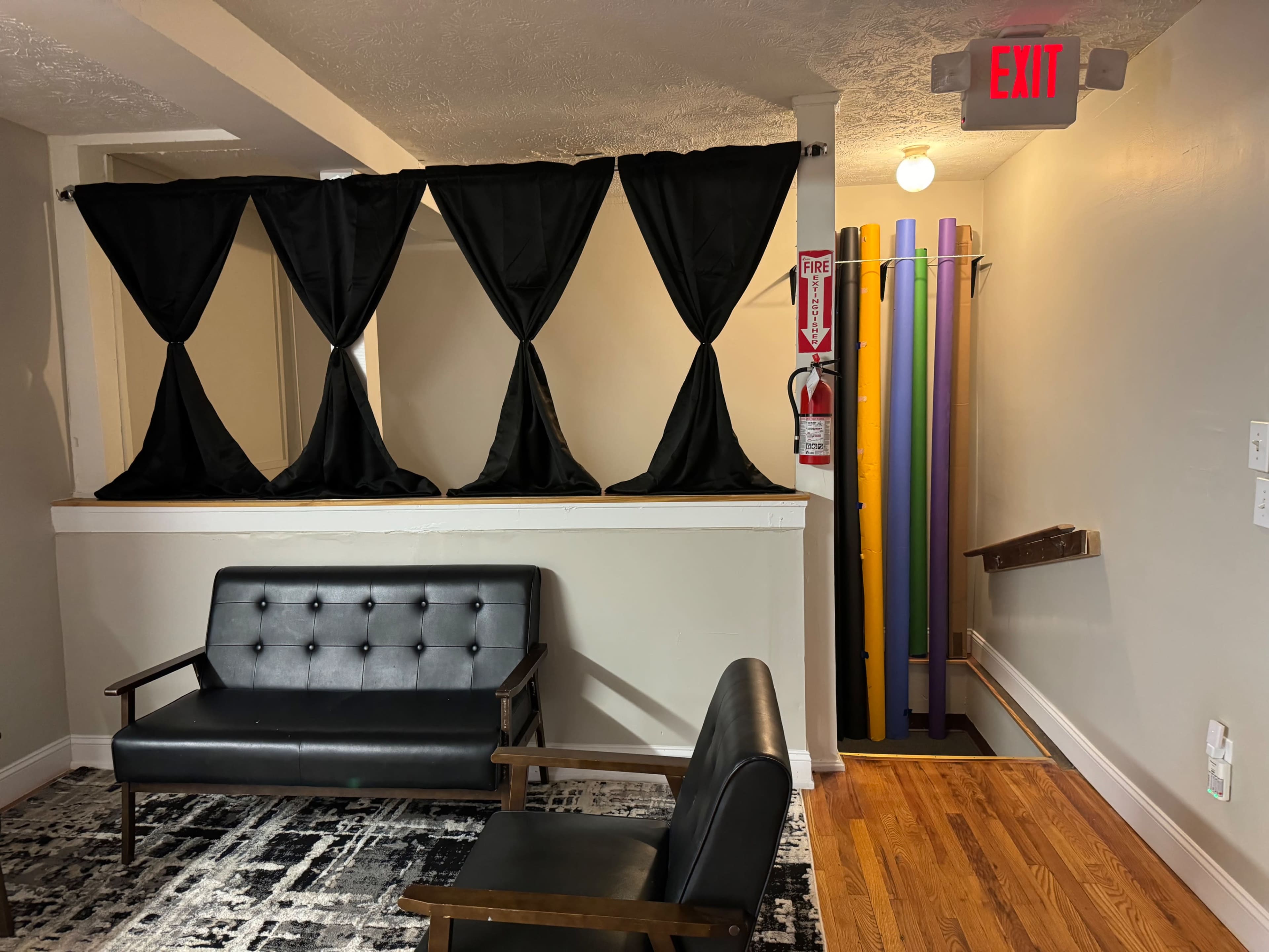 A room with a black sofa in front of a partition of black curtains, next to a hallway featuring colorful vertical poles and an exit sign.