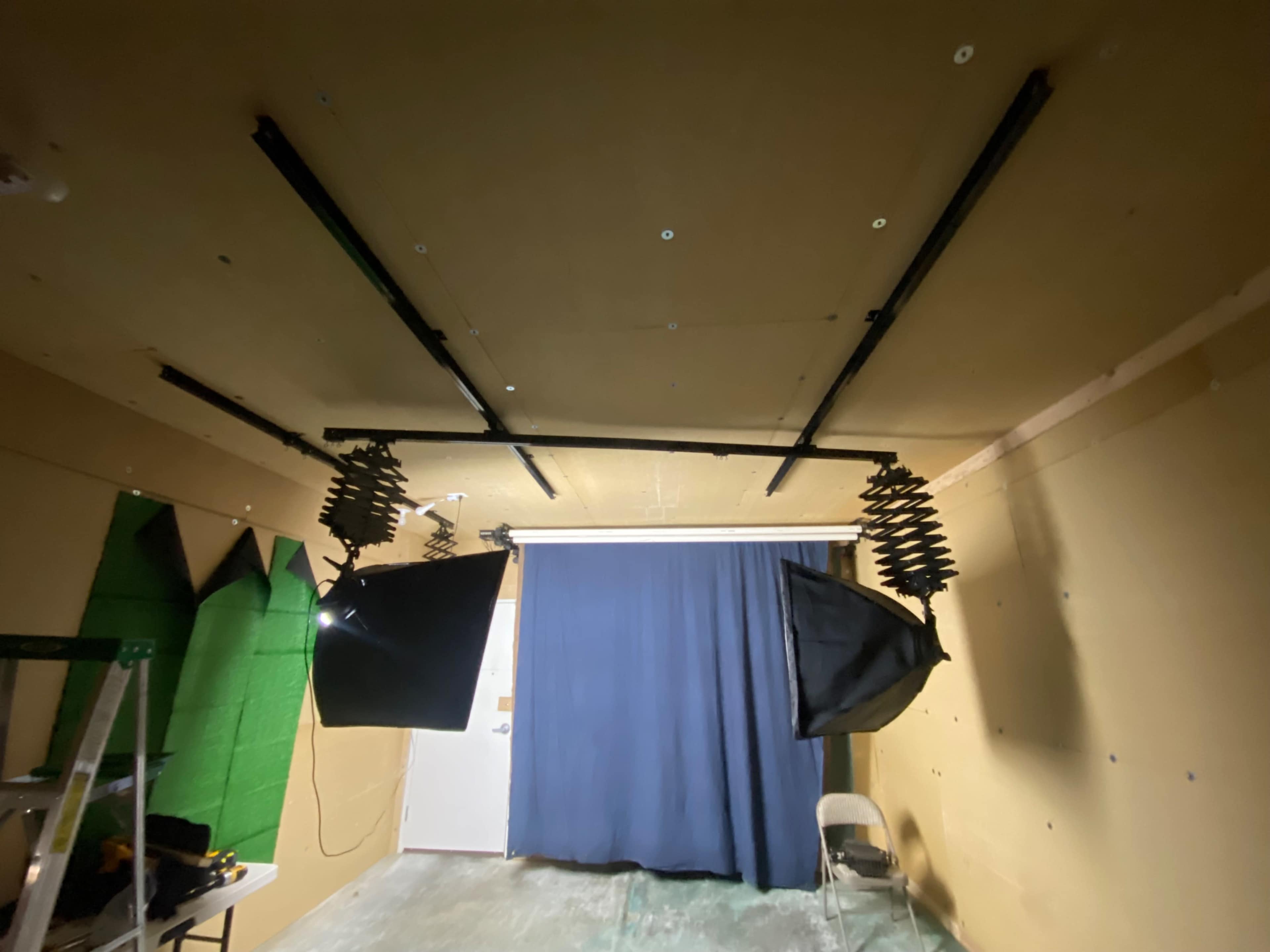 The image shows a room with a ceiling mounted lighting setup, a closed blue curtain, and green fabric pieces on one wall, alongside a ladder and a chair.