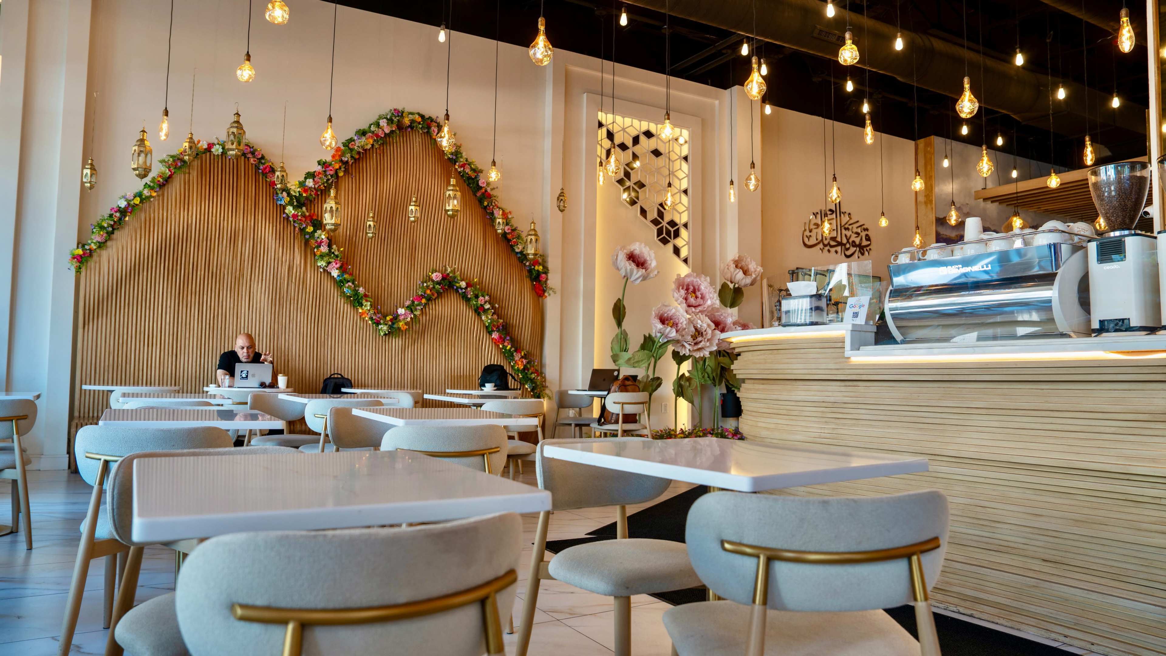 The image shows a modern café interior with floral decorations on a wooden wall and a counter topped with a coffee machine and pastries.