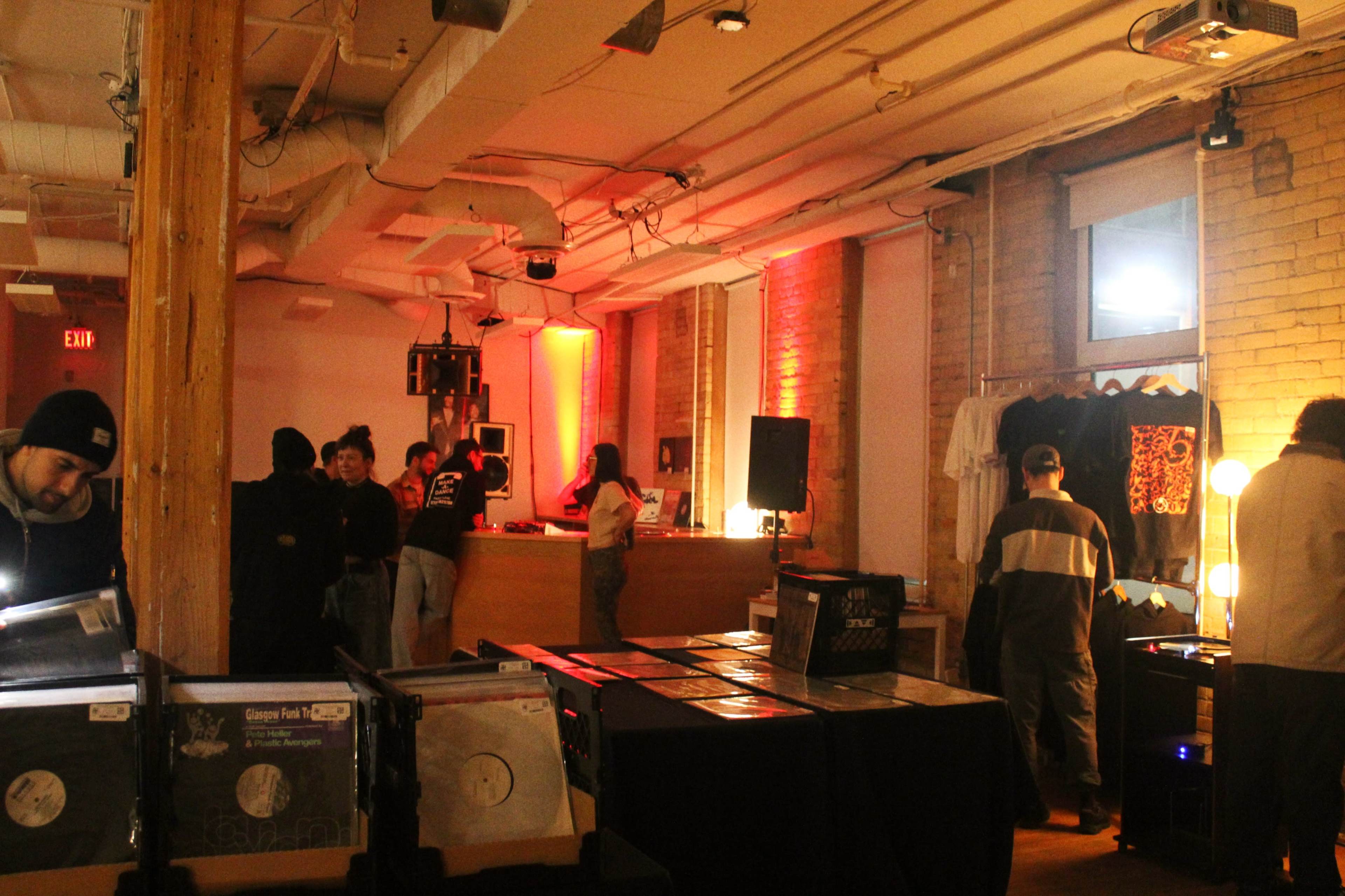 A group of people interacts in a dimly lit room lined with wooden beams, where vinyl records are displayed on tables and a DJ setup is visible.