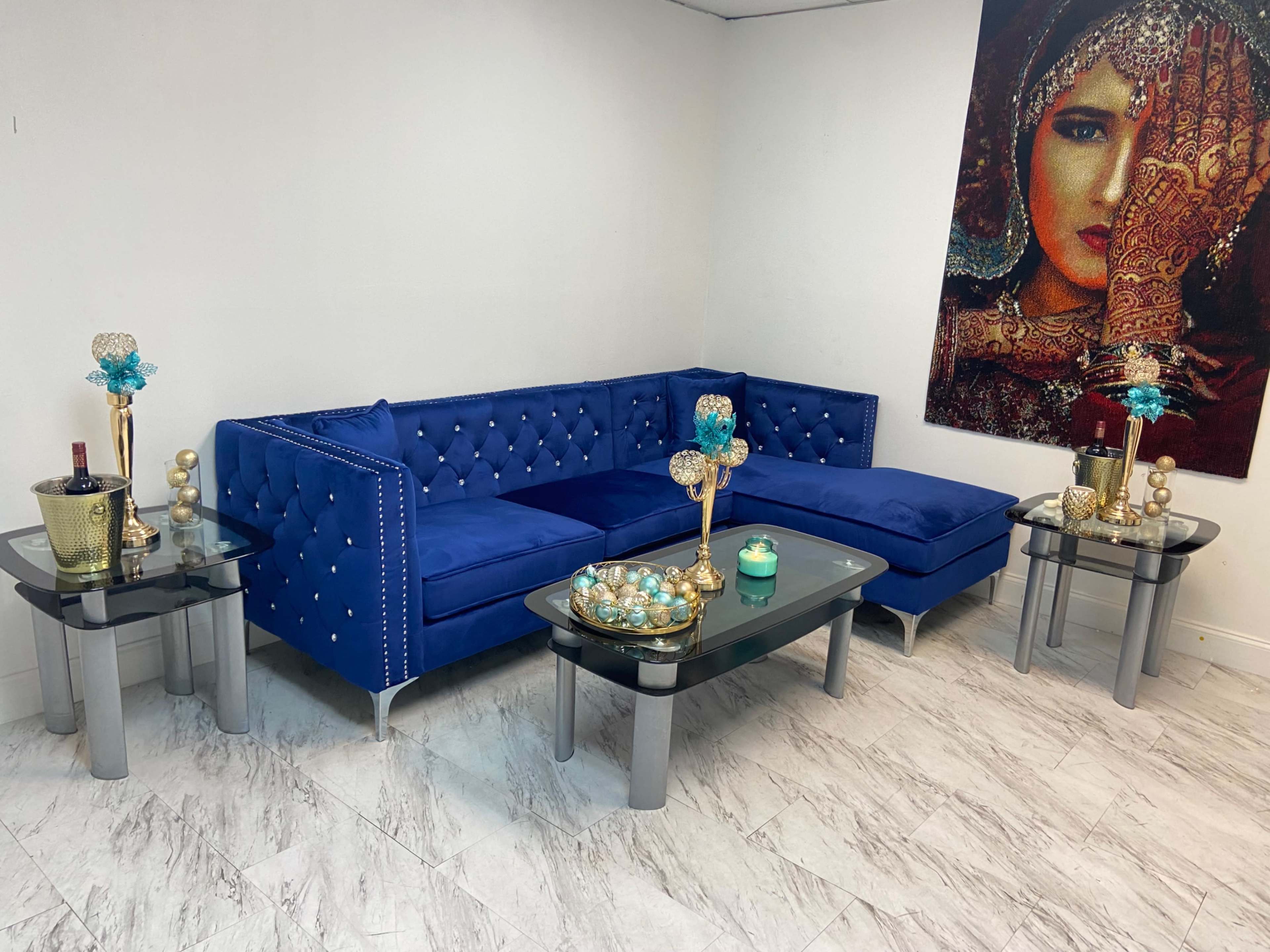 The image shows a blue velvet sectional couch accompanied by two glass side tables, each featuring decorative items, set against a white wall adorned with a framed artwork of a woman.