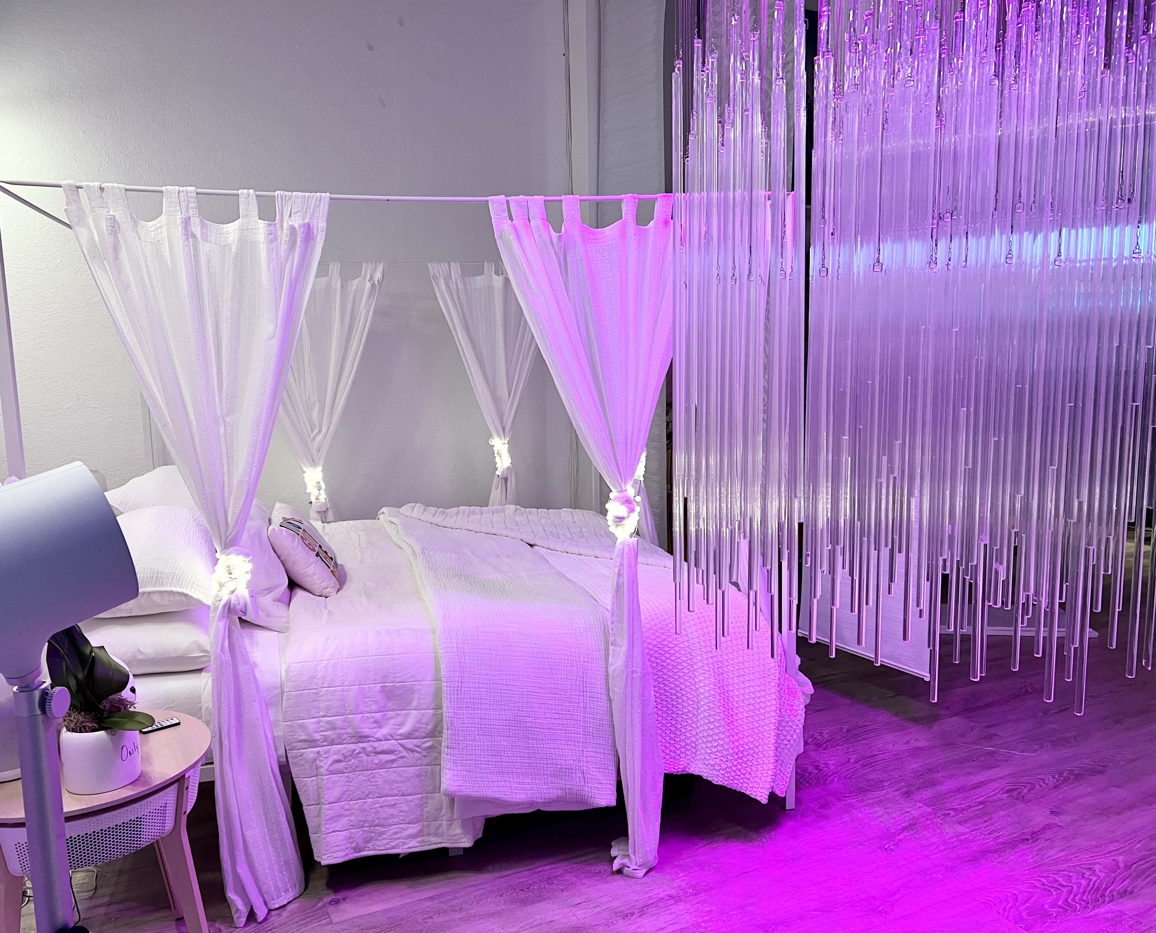 A bedroom features a bed with a white blanket and sheer curtains draped overhead, illuminated by soft purple lighting from hanging decor.