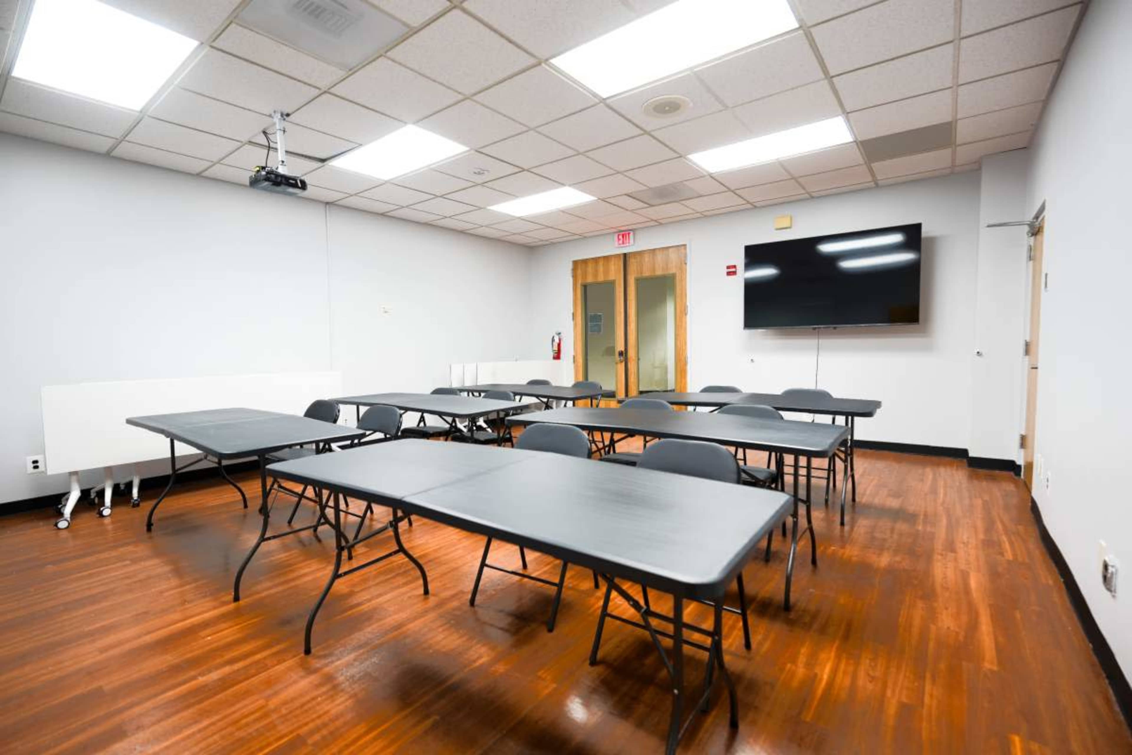 The image shows a meeting room with several black folding tables arranged in rows, a wall-mounted television, and a projector hanging from the ceiling.