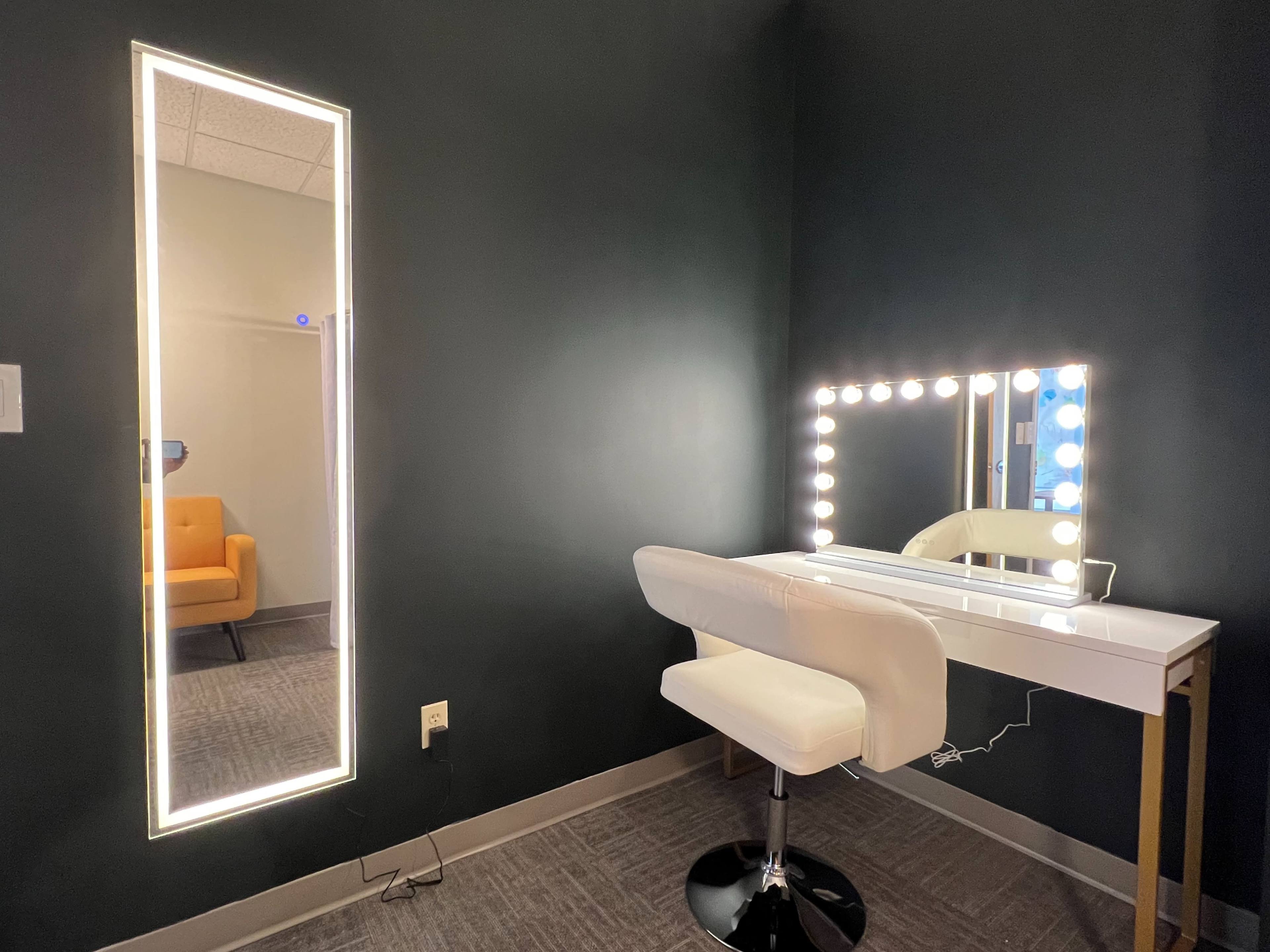 A dressing area featuring a illuminated vanity mirror and a backlit full-length mirror, both against a dark wall, with a white vanity table and a stool adjacent to them.