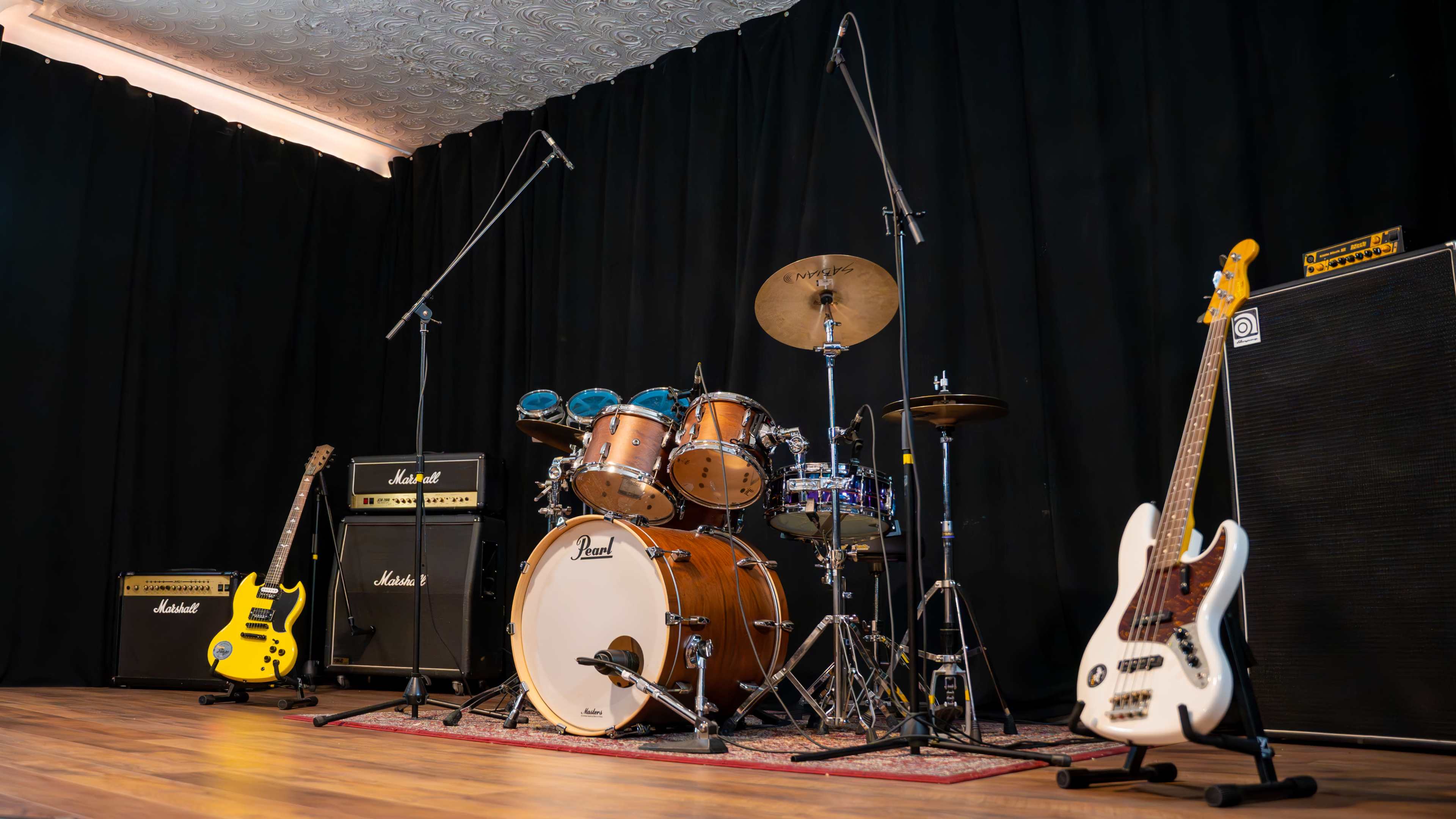A drum set with cymbals, two electric guitars, amplifiers, and a microphone stand are arranged on a stage with a dark backdrop.