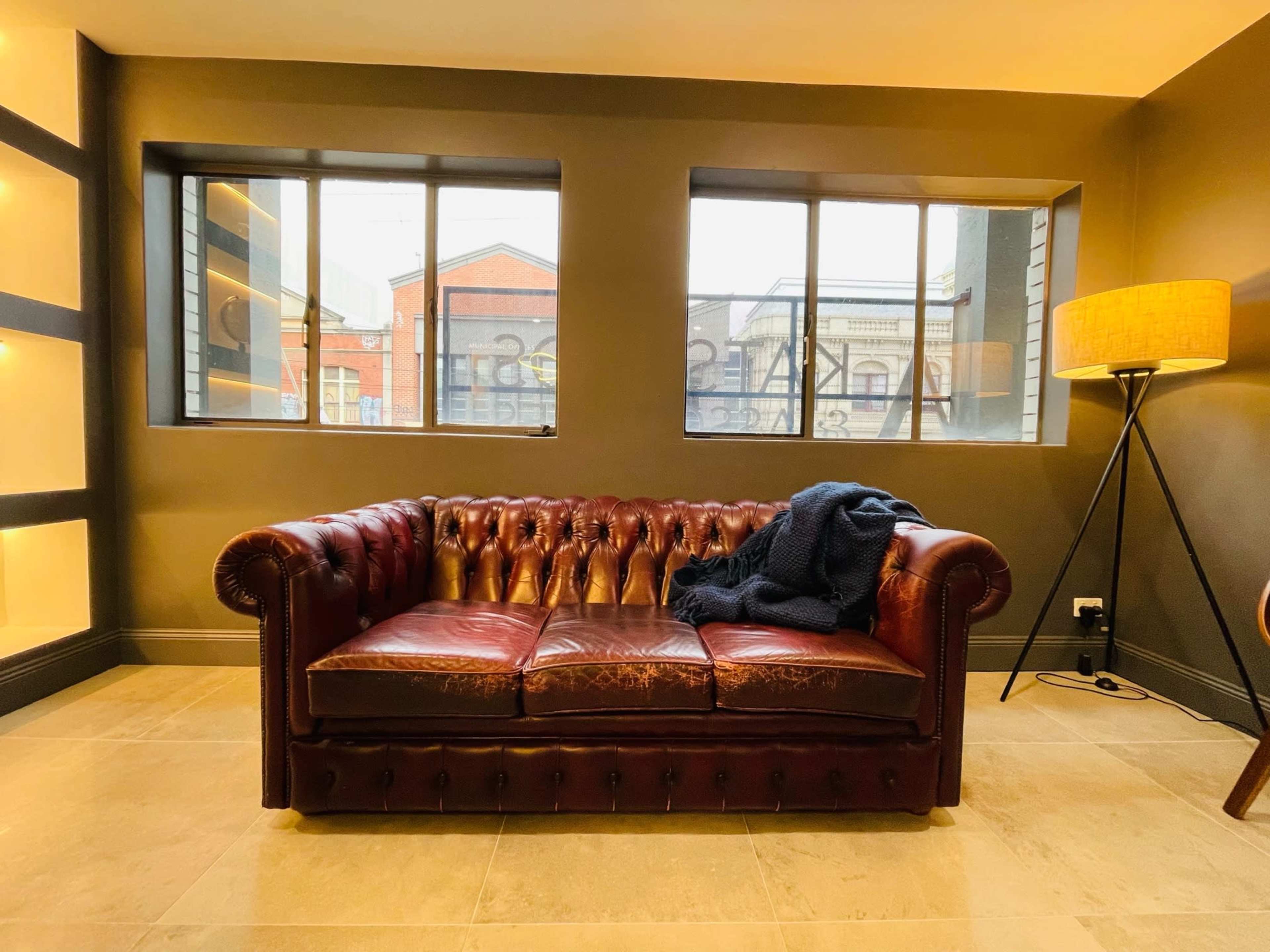 The image shows a brown leather Chesterfield sofa with a dark throw draped over it, positioned against a wall with large windows that allow natural light into the room.