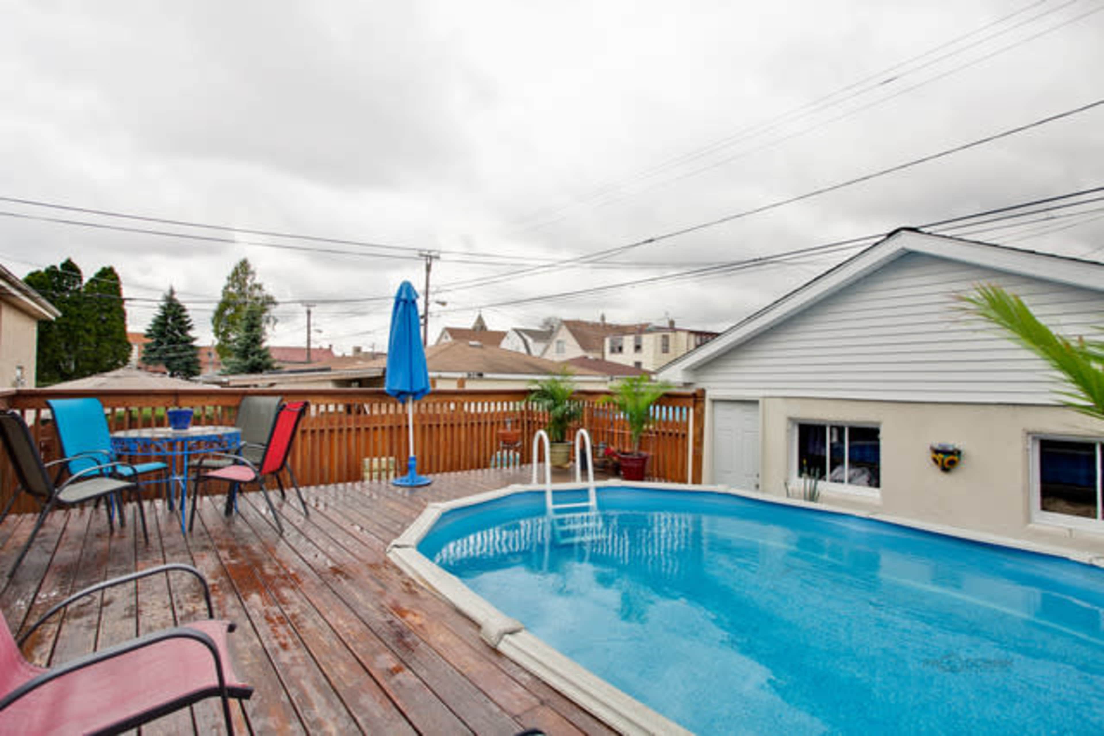 The image shows a wooden deck with a blue pool, surrounded by chairs and a table, and a house in the background under a cloudy sky.