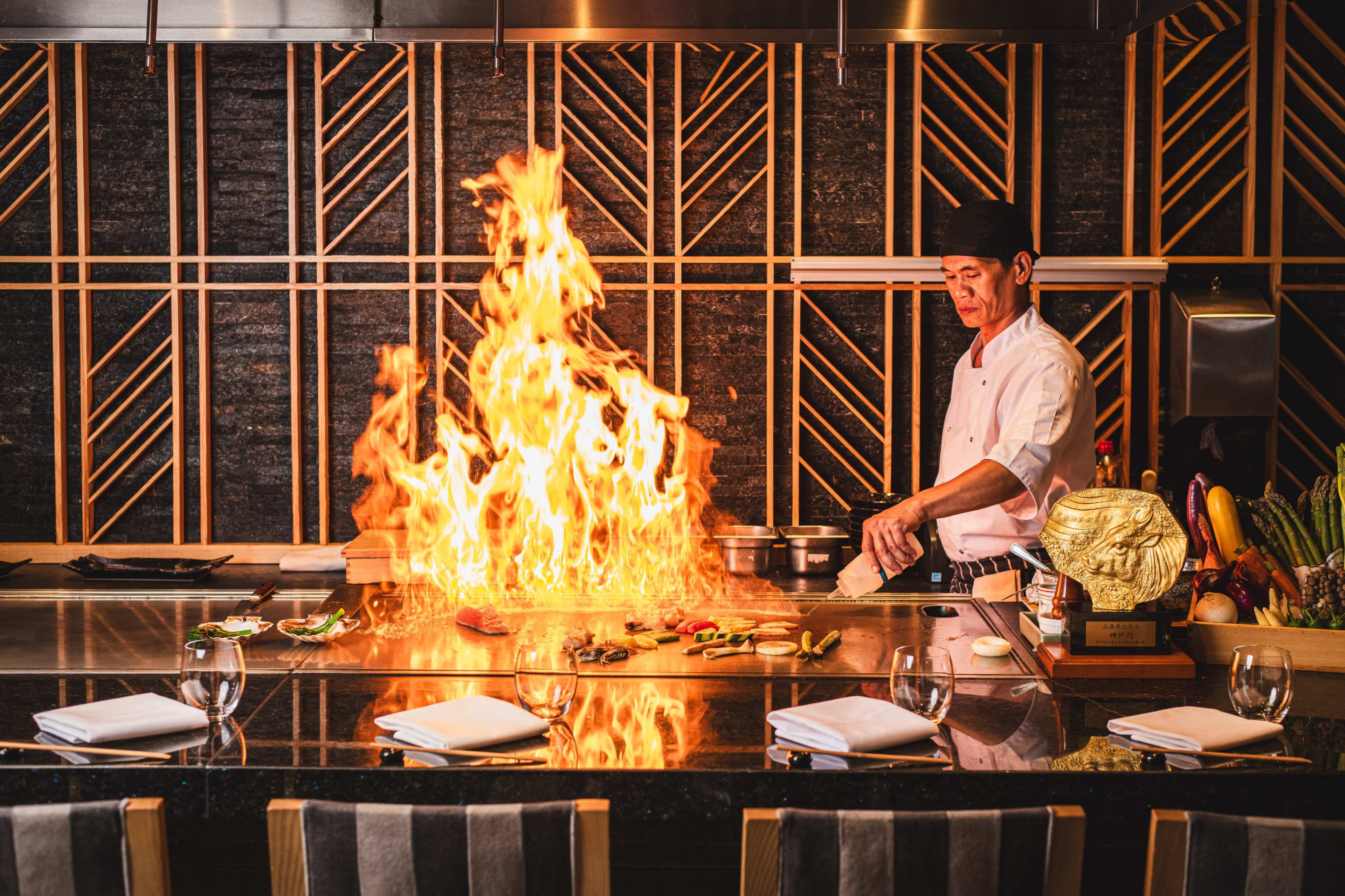 A chef at a teppanyaki grill prepares food as flames rise dramatically from the cooking surface.