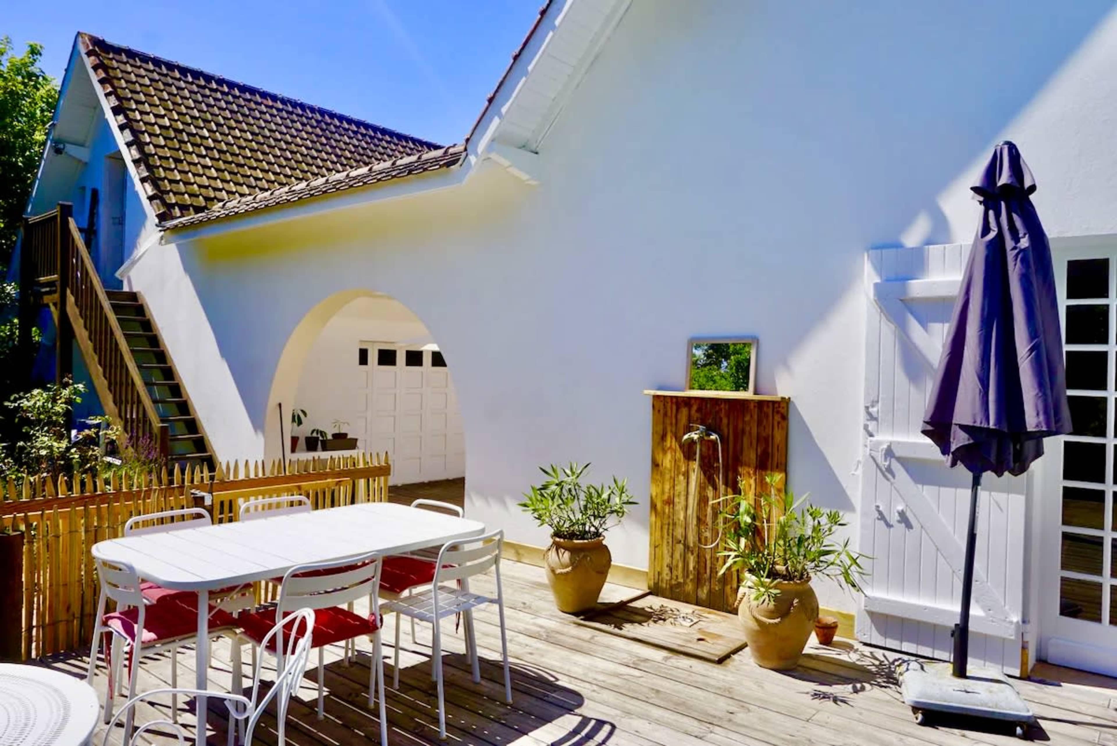 The image features a bright outdoor patio area with a dining table, potted plants, an umbrella, and an outdoor shower beside a house.
