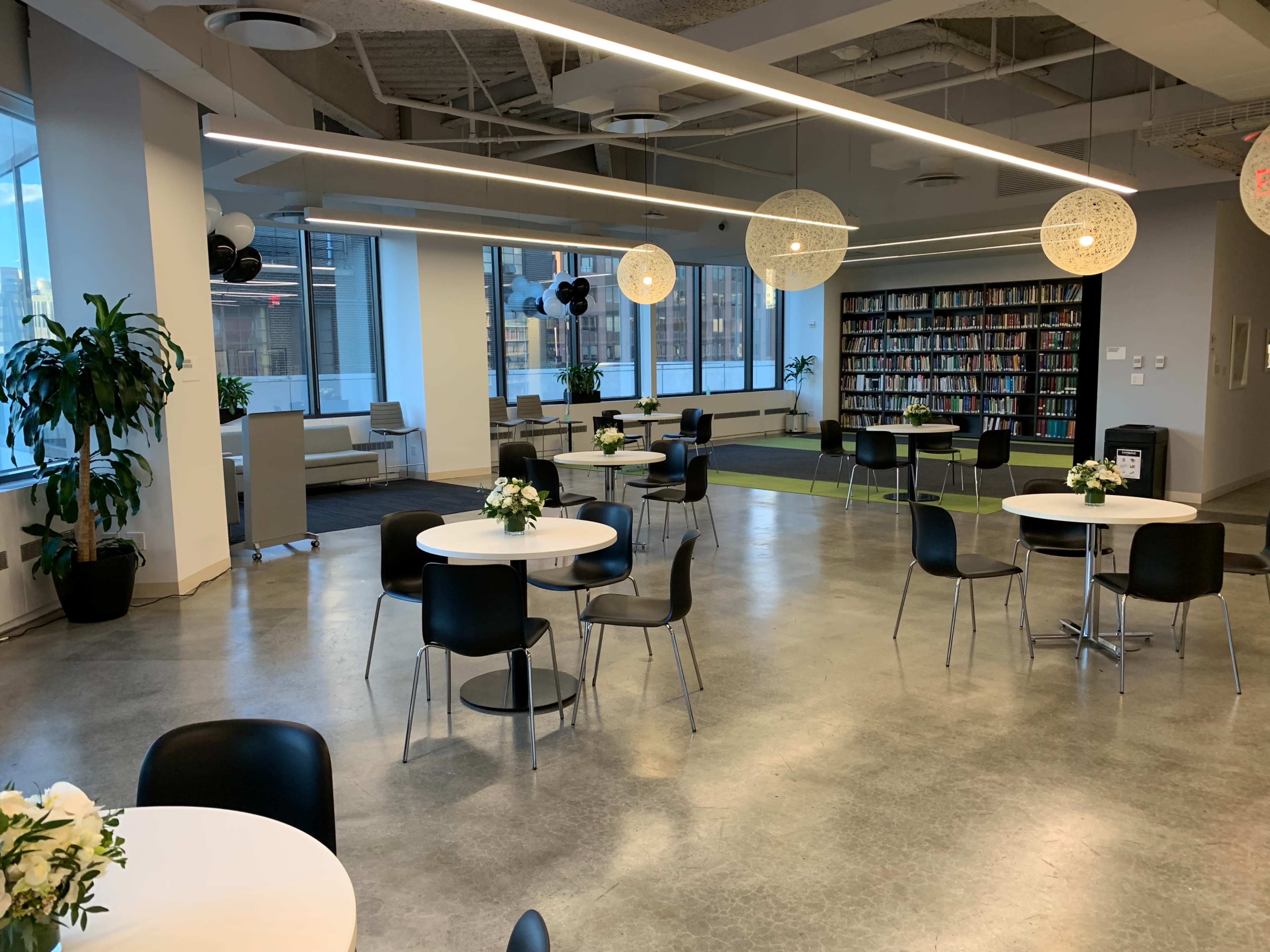 A modern, open space with several round tables and chairs, decorated with small floral arrangements, and large windows illuminating the area.