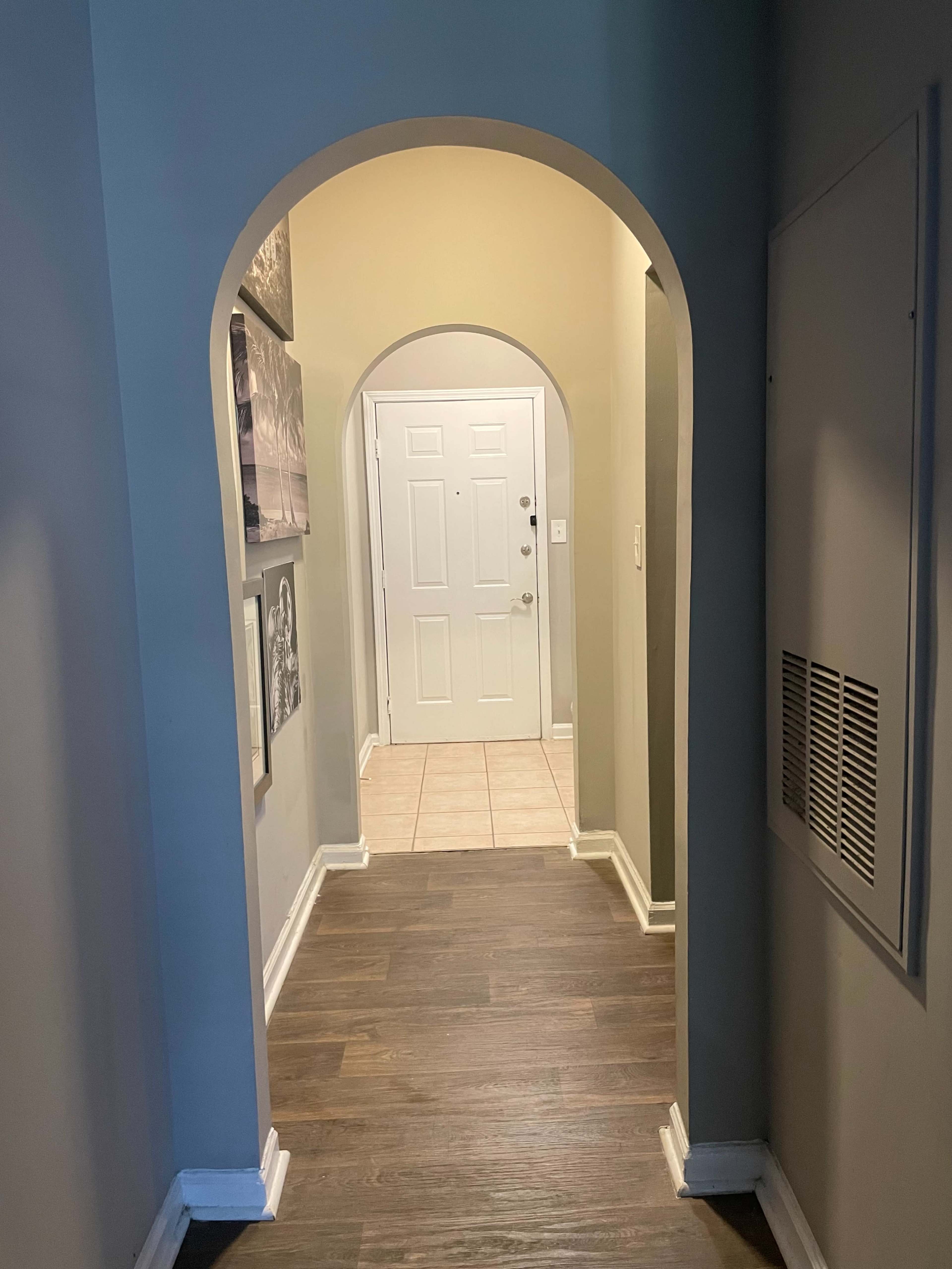 The image shows a narrow hallway with an arched entrance leading to a door at the end and framed photographs on the wall.