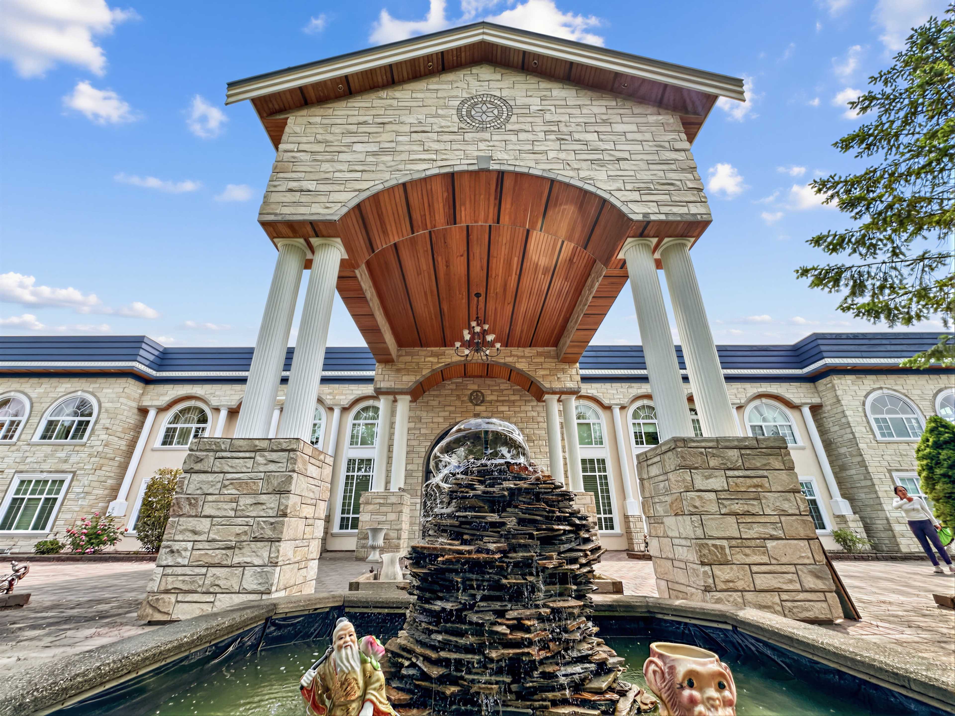 The image shows a large stone structure with a raised roof and decorative columns, featuring a fountain with sculptures in the foreground and a well-maintained garden in the background.