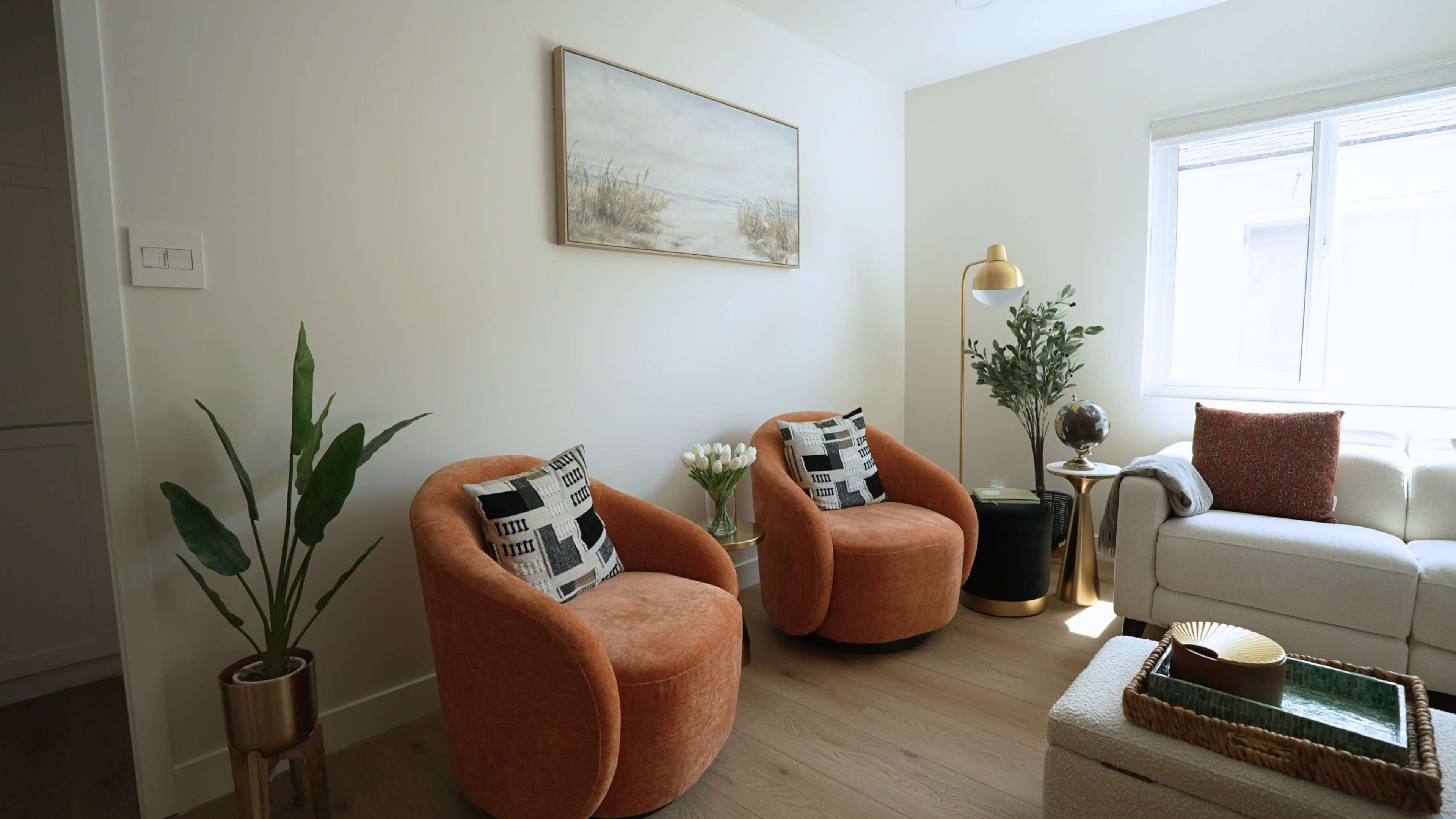 The scene features a cozy living room with two orange, curved chairs, a white sofa, a small table, a floor lamp, plants, and a painting on the wall.