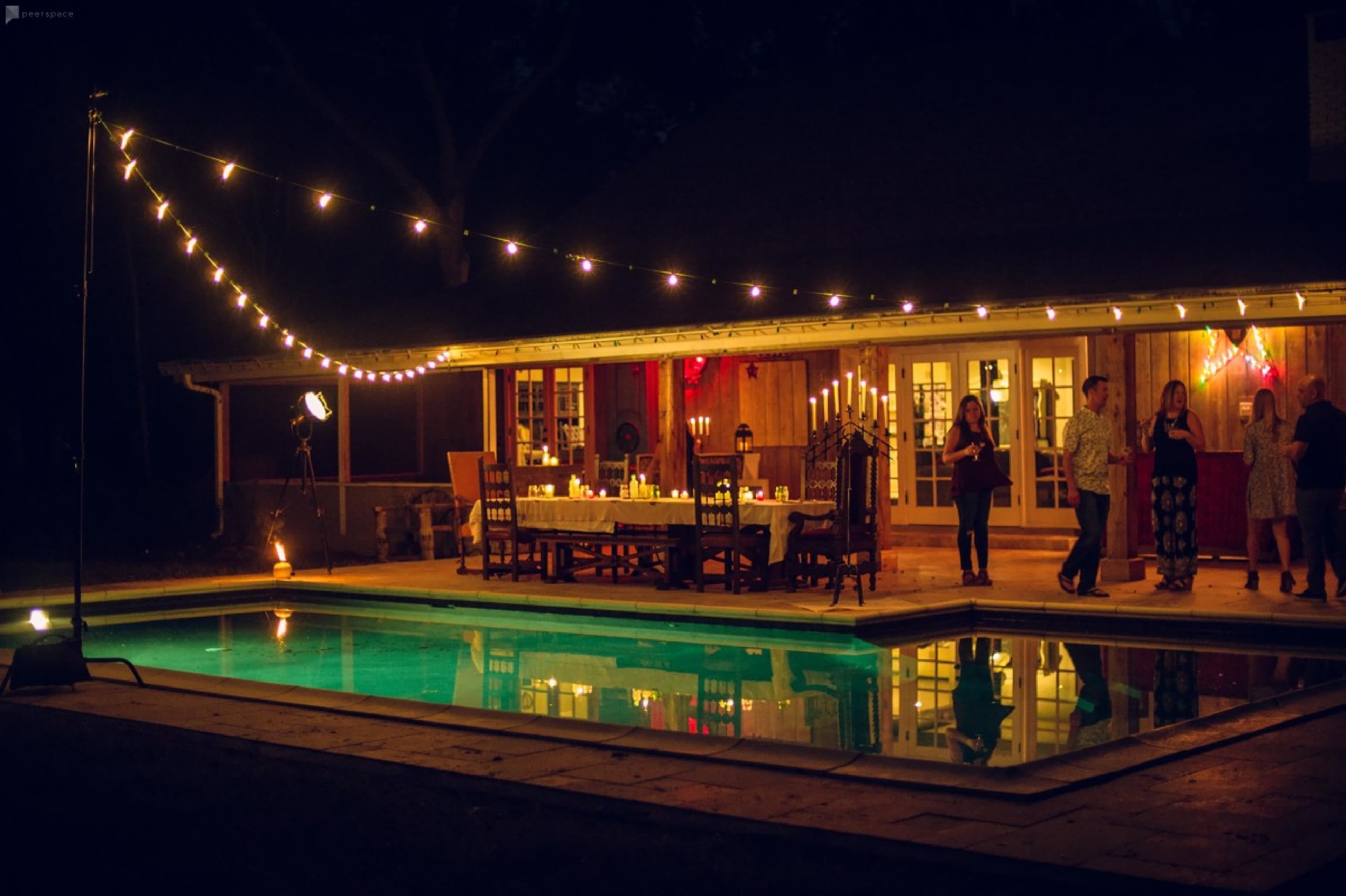 A beautifully set outdoor dining area is illuminated by string lights beside a swimming pool at night.