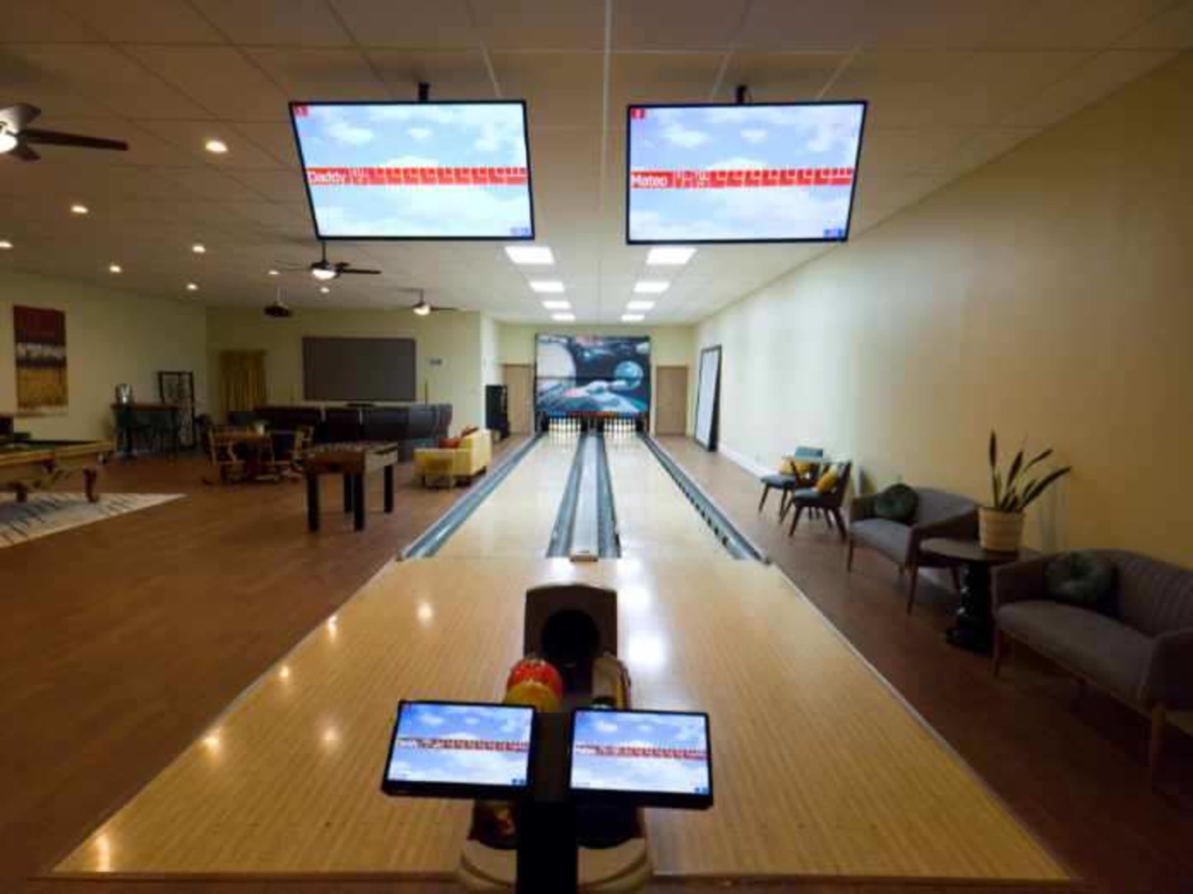An indoor bowling alley with three large screens displaying scores, and a smooth wooden lane in the center, flanked by seating areas.