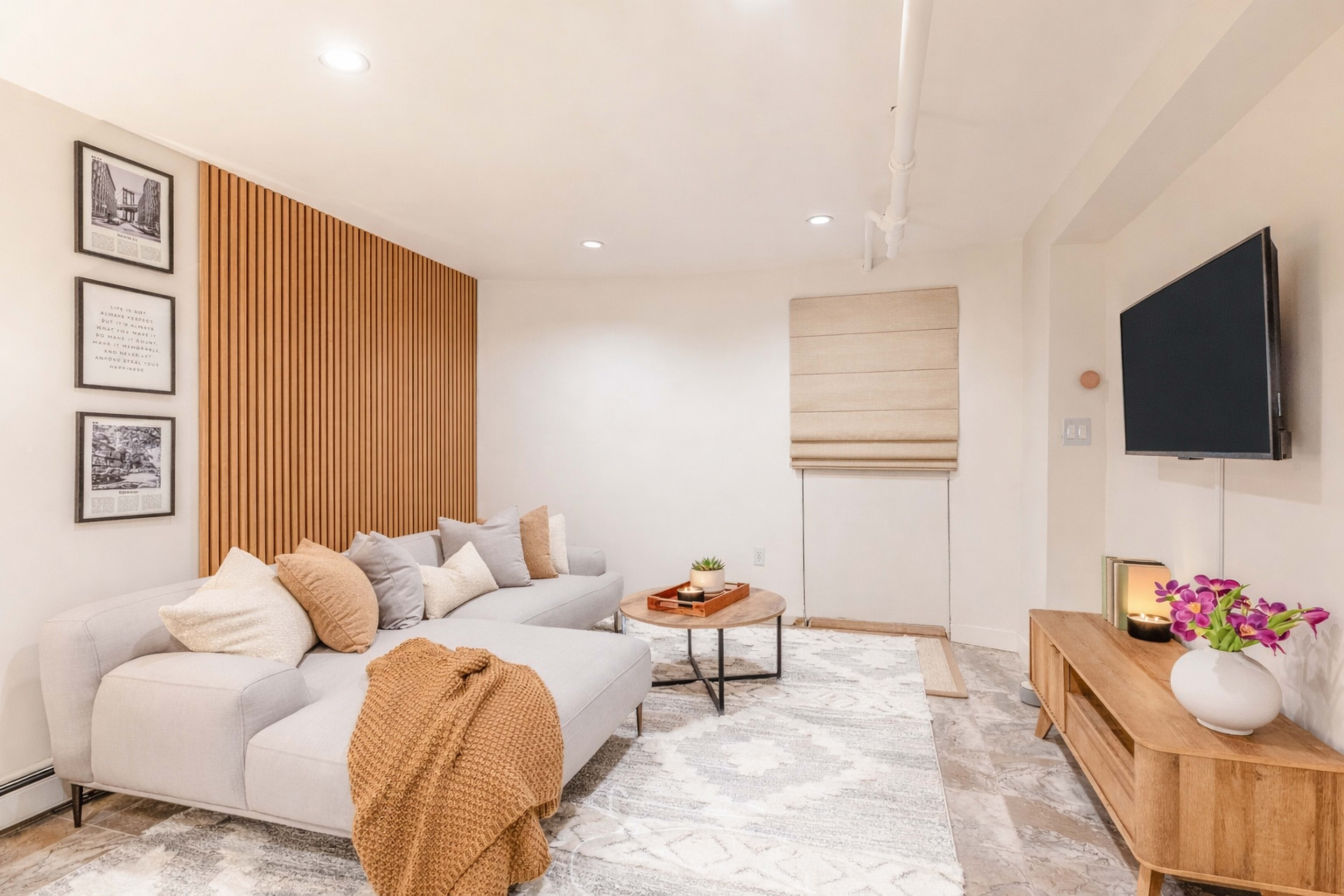 The image shows a cozy living room with a light-colored sectional sofa, decorative pillows, a wooden coffee table, and a wall-mounted television, complemented by framed artworks and a window with a beige roller shade.