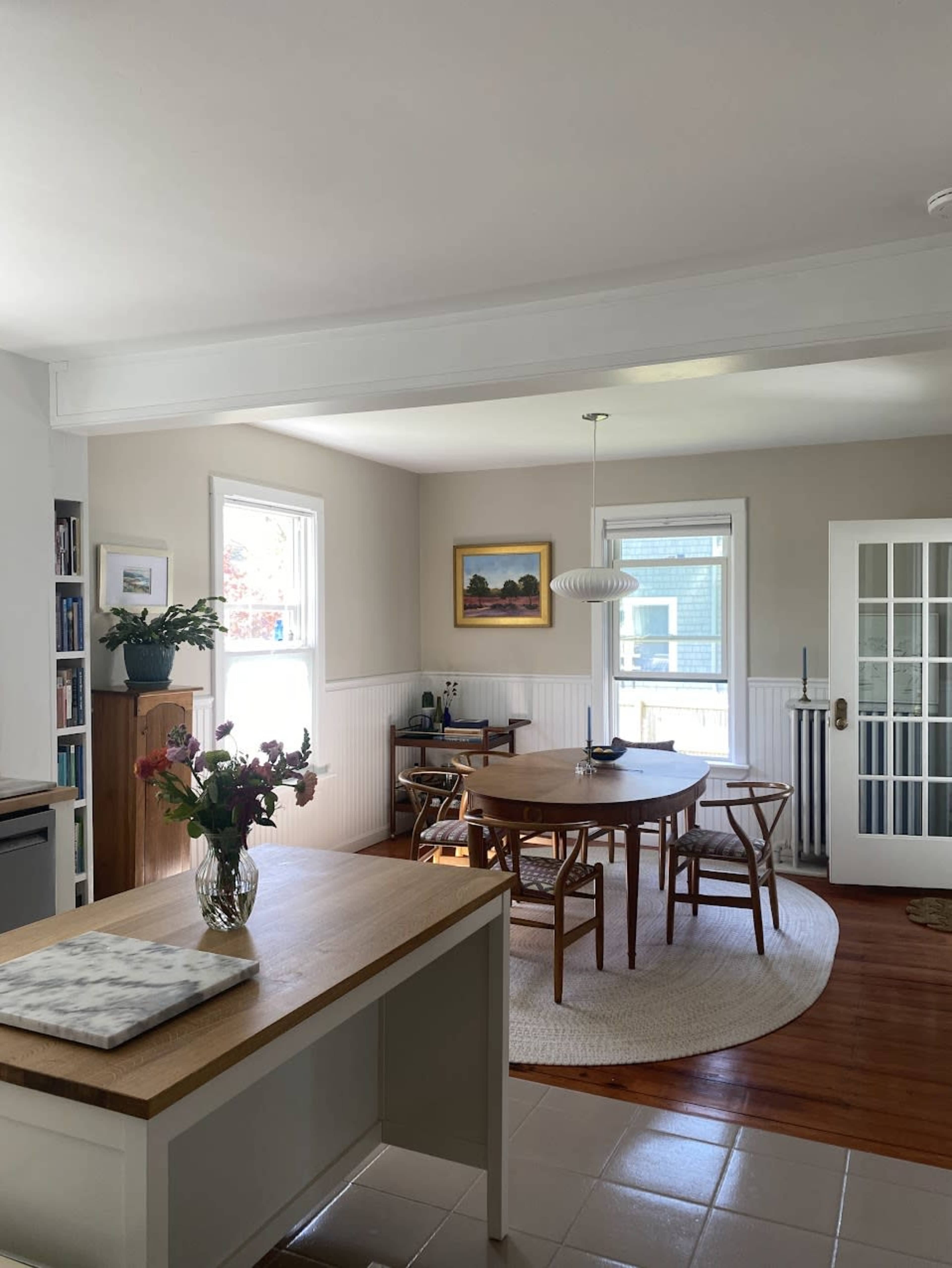 A well-lit kitchen and dining area features a wooden table surrounded by chairs, a vase of flowers on the counter, and large windows providing natural light.