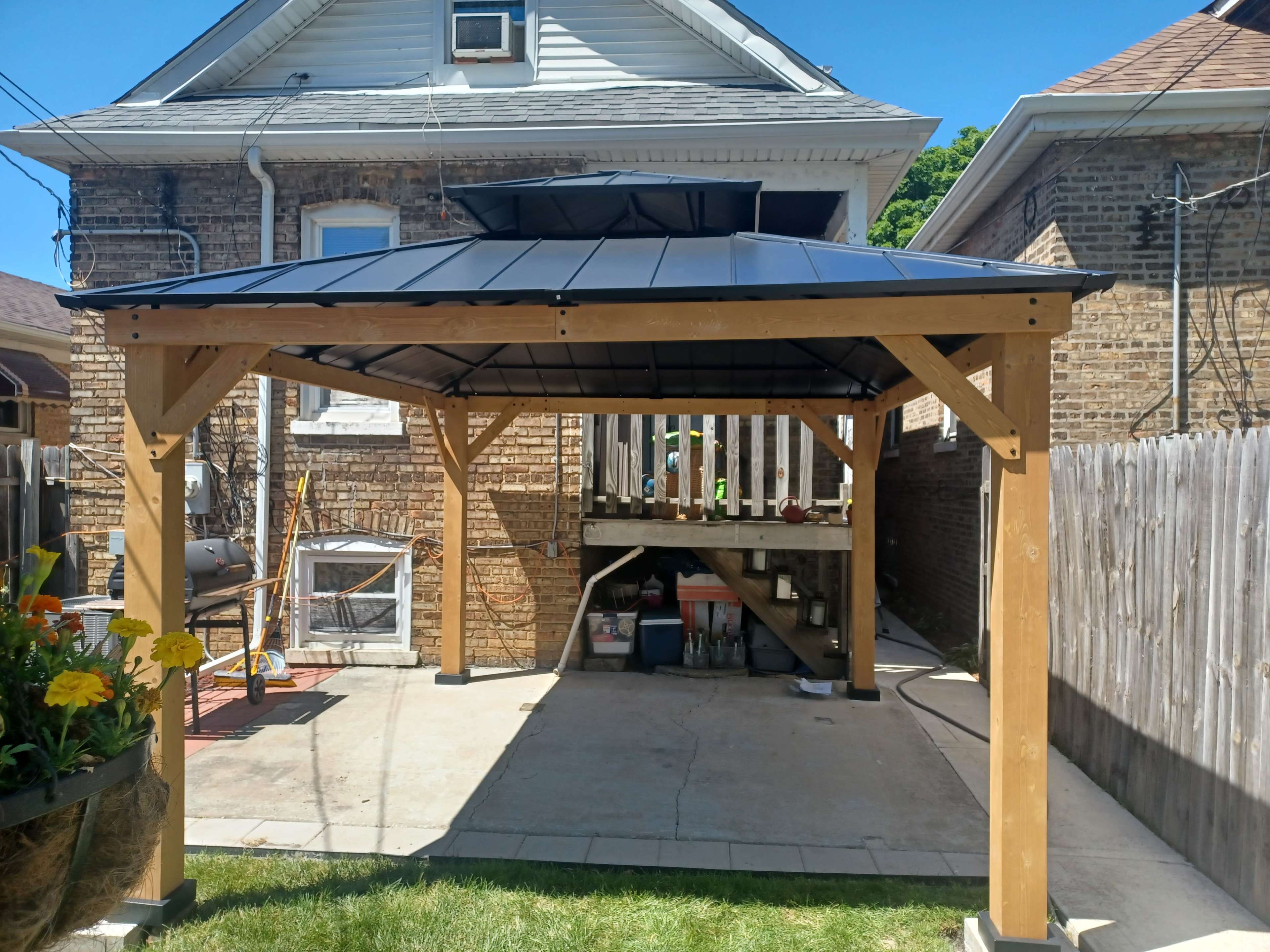 A wooden gazebo with a black roof, situated on a concrete surface next to a house and a wooden fence.