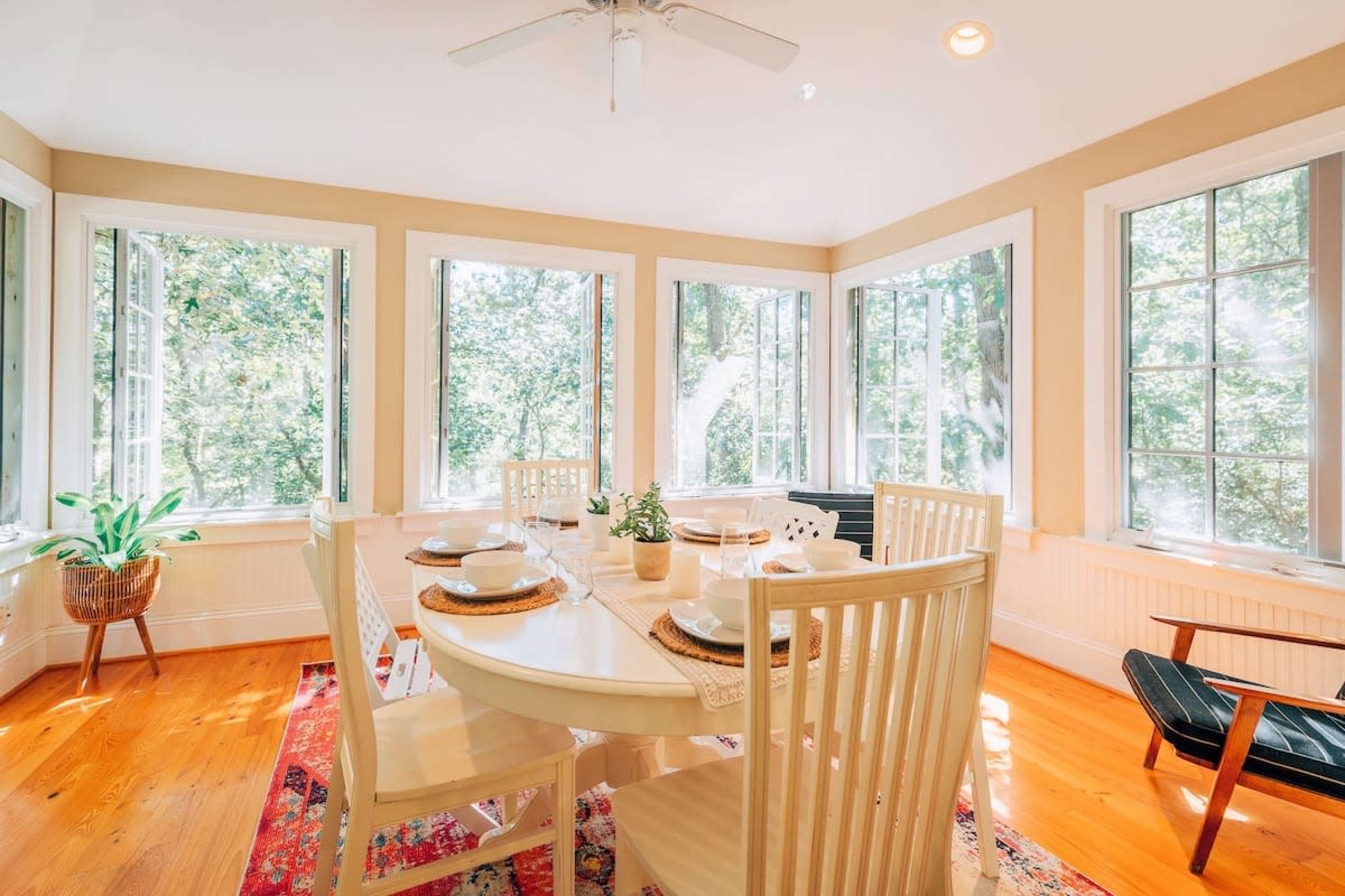 A bright dining area with a round table set for a meal, surrounded by large windows offering views of trees outside.