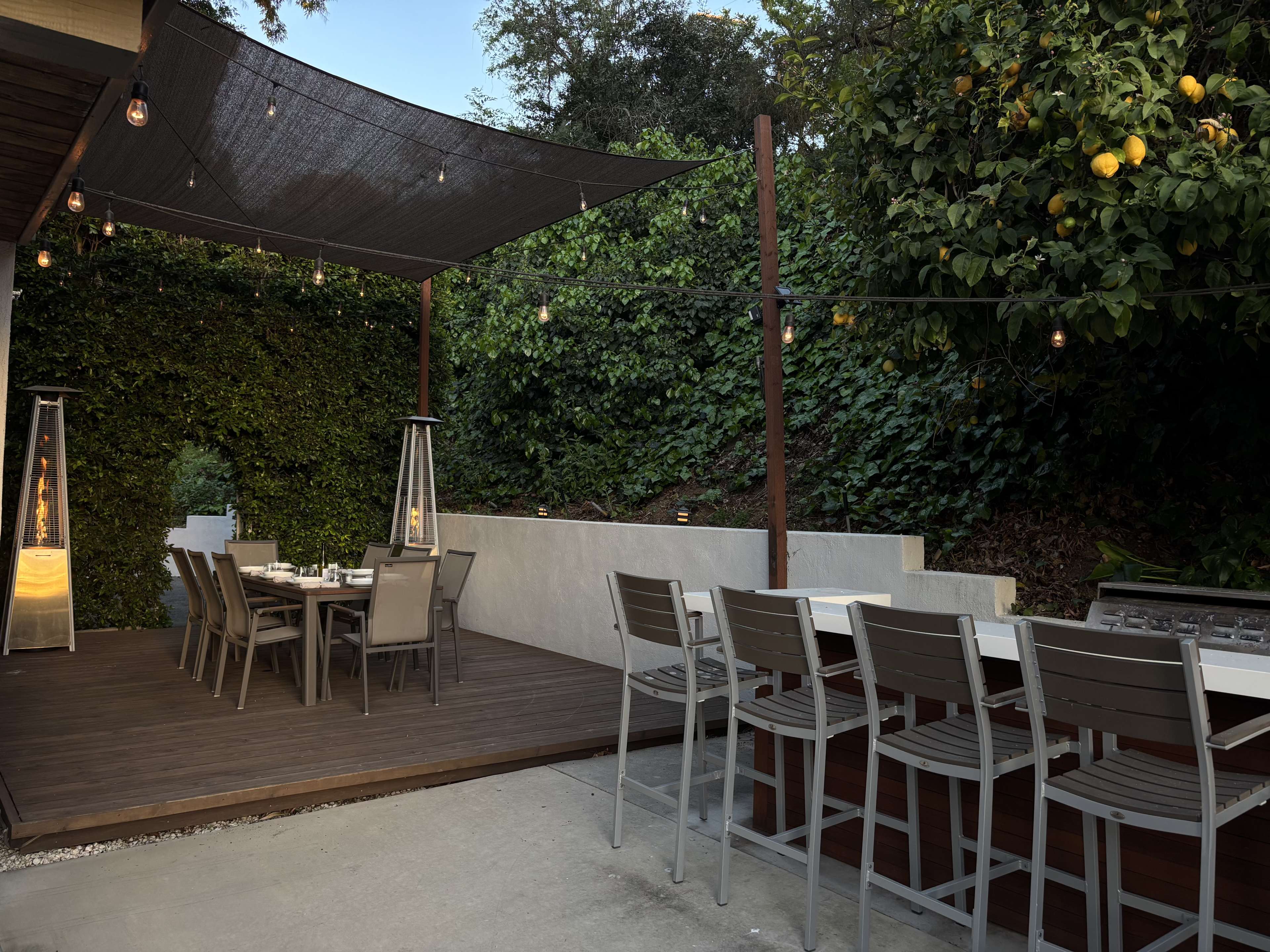 The image shows an outdoor dining area with a wooden deck, a large table with chairs, and a shaded area surrounded by greenery and string lights.