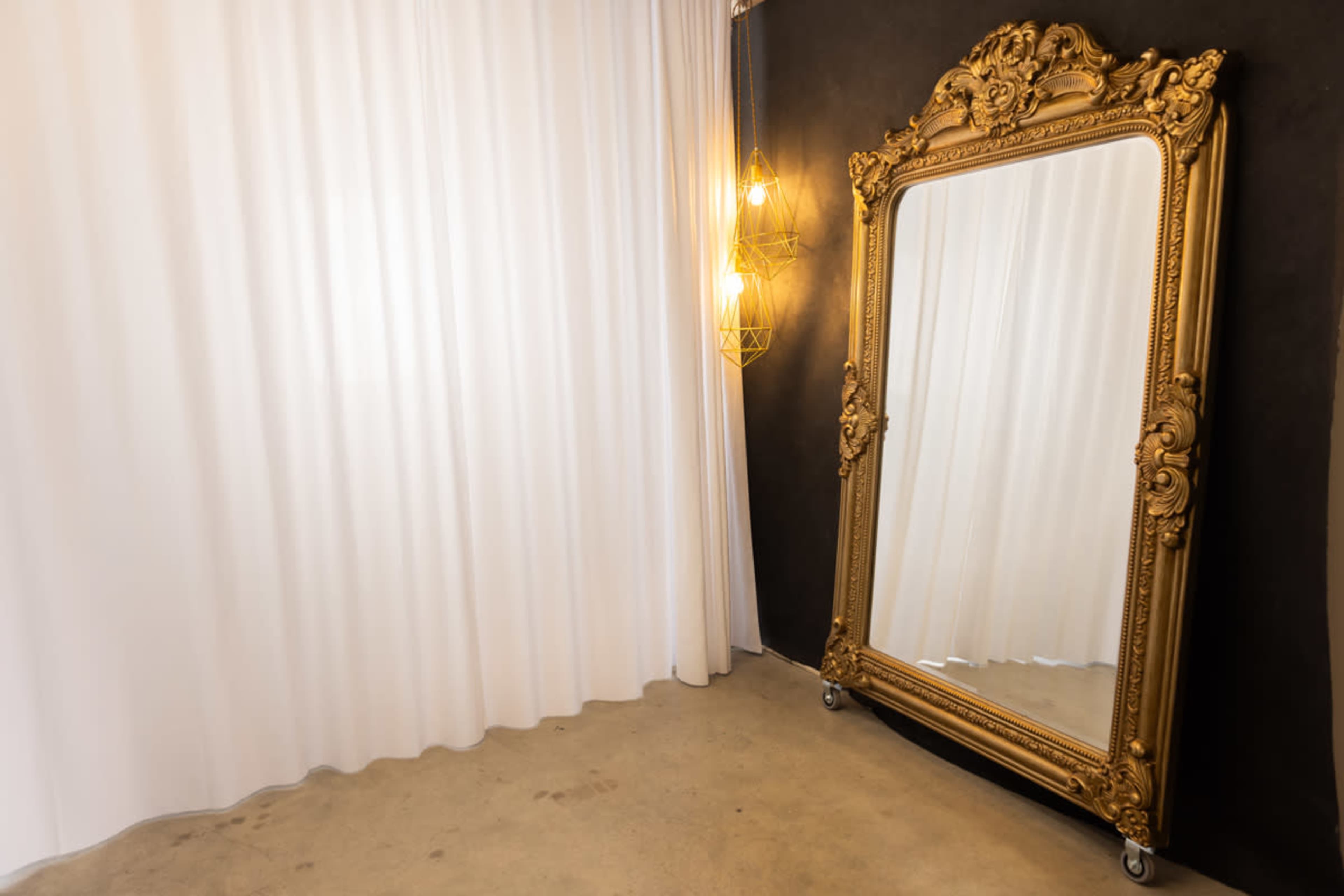 A large ornate gold mirror is positioned next to a white curtain and illuminated by pendant lighting on a dark wall.