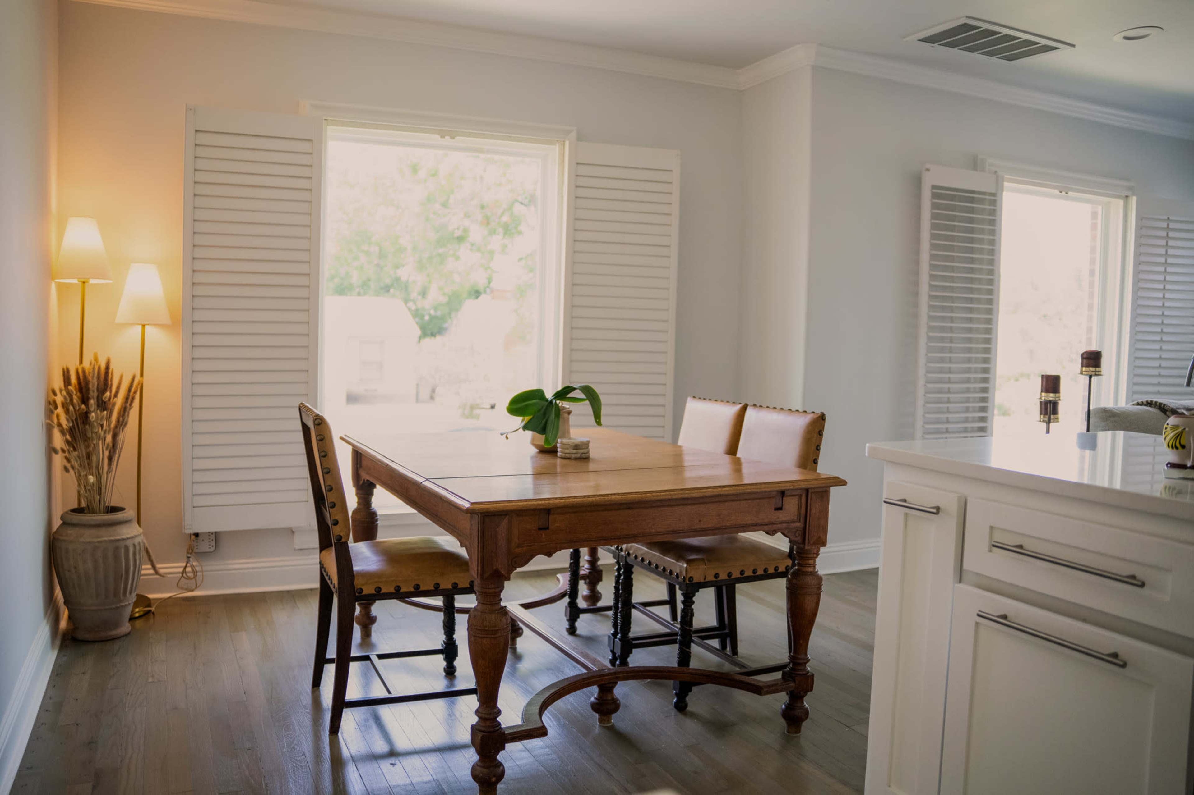 A wooden dining table with two chairs is set in a well-lit room featuring large windows and simple decor.
