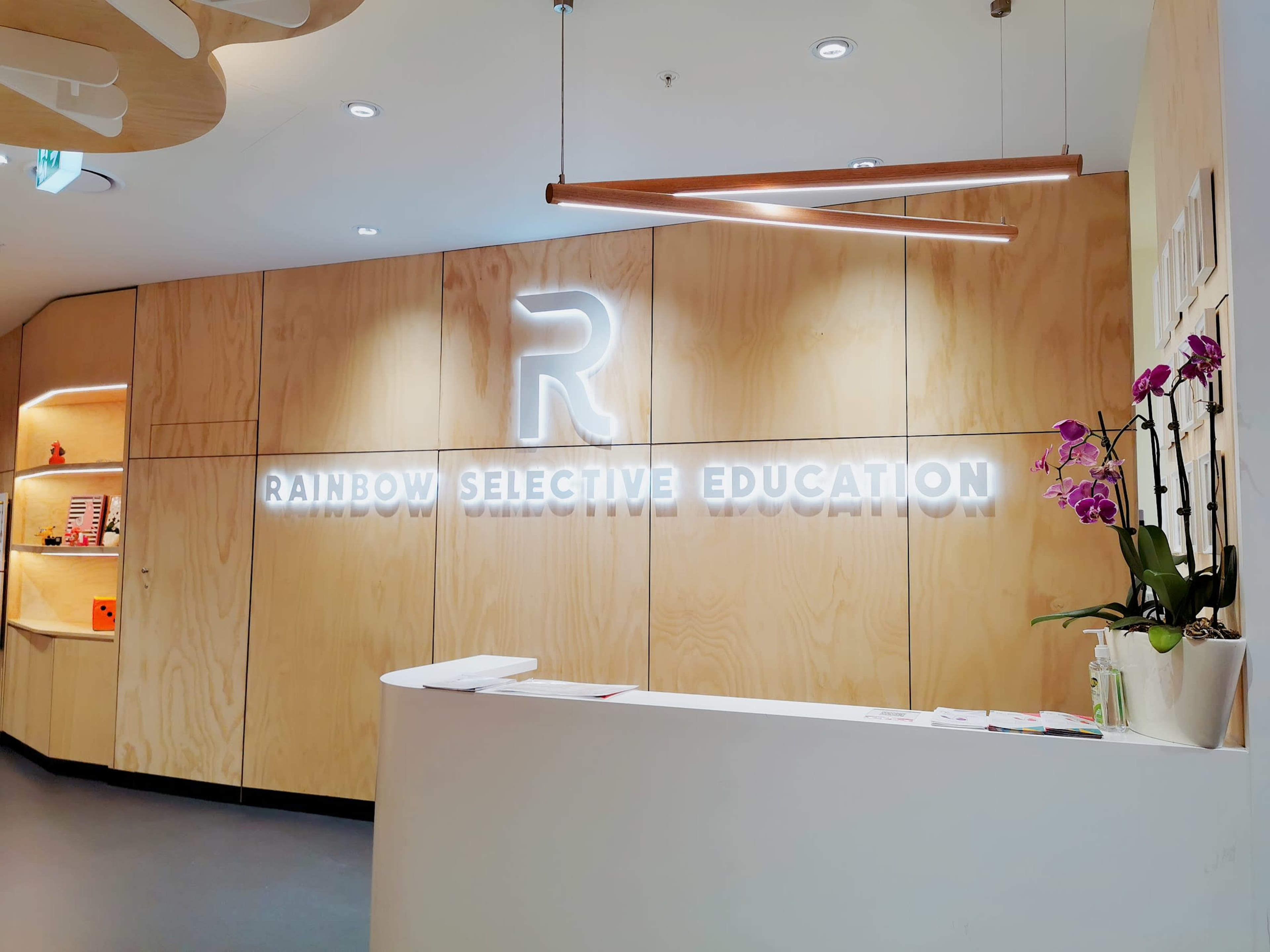 The image shows the reception area of an educational center with a wooden wall featuring a glowing "RAINBOW SELECTIVE EDUCATION" sign and a curved front desk.
