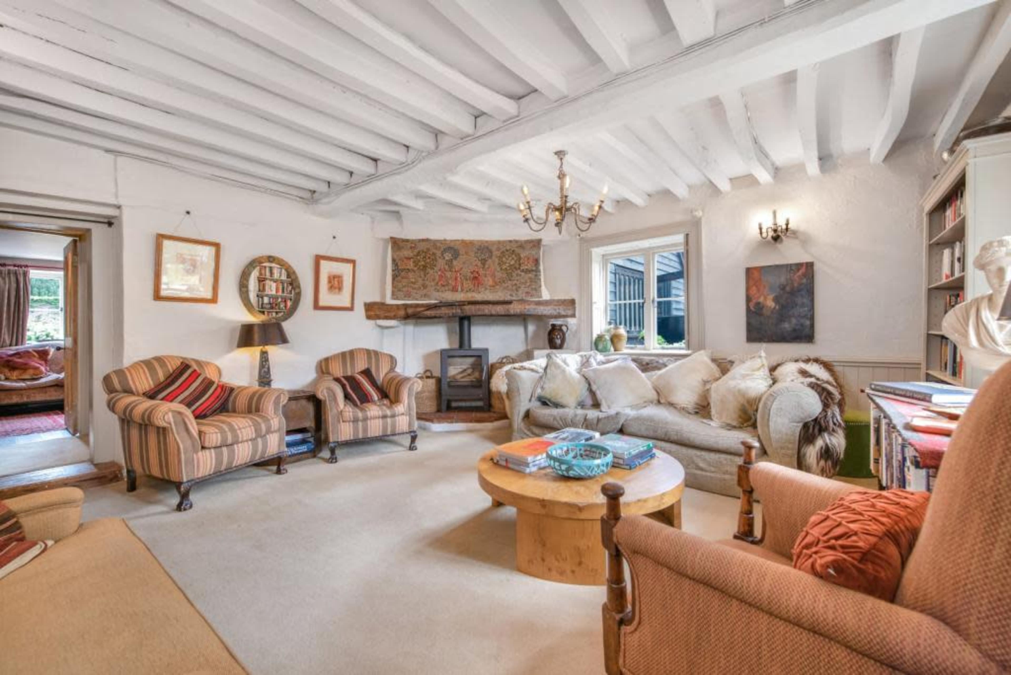 The image shows a cozy living room with two striped armchairs, a large sofa, a wooden coffee table, a fireplace, and exposed wooden beams on the ceiling.