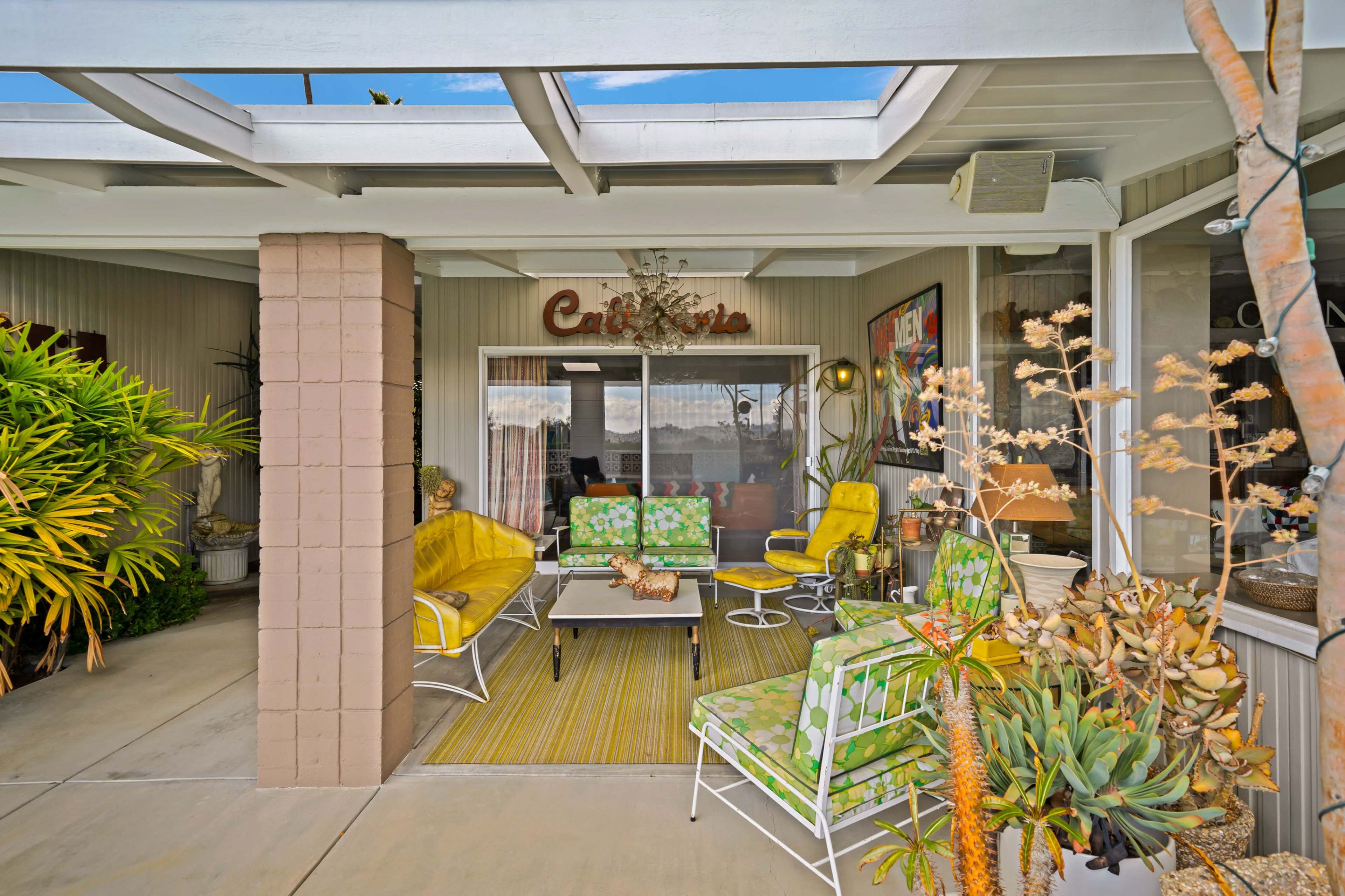 A covered patio features green and yellow furniture surrounded by potted plants and decorative elements.
