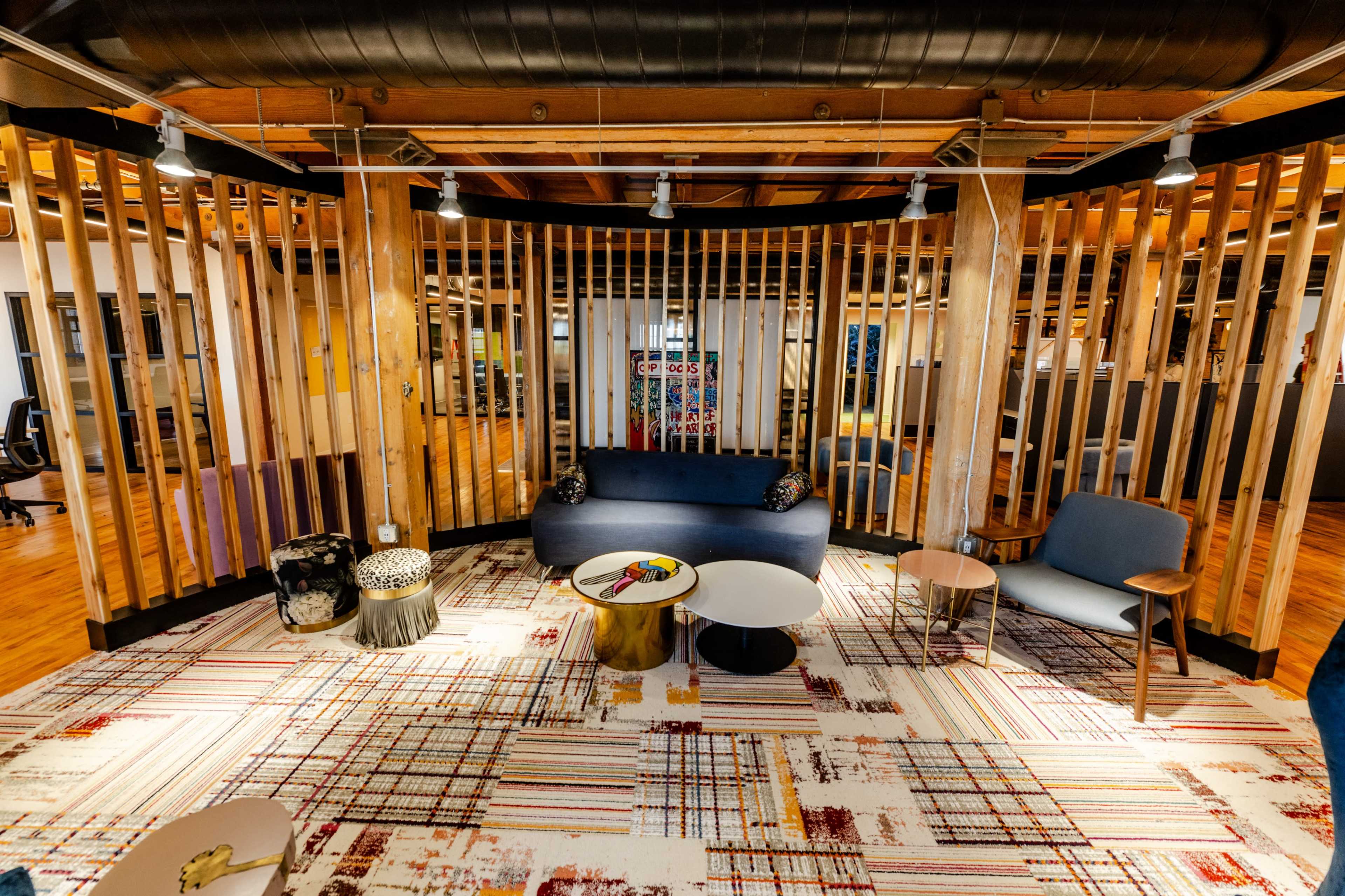 The image shows a modern office space featuring wooden slats that create a semi-enclosed seating area, furnished with a navy sofa, various chairs, and round tables on a patterned rug.