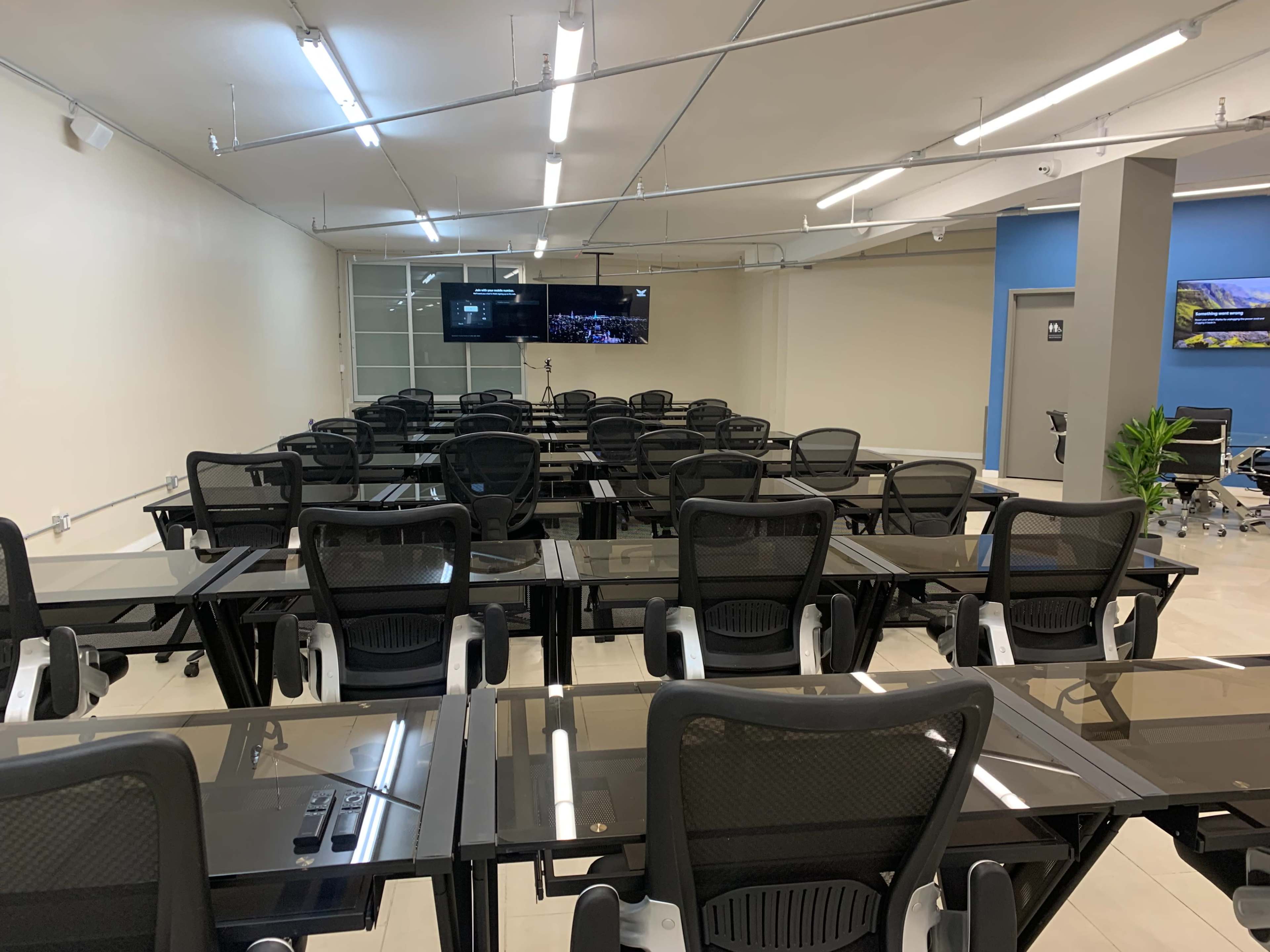 A classroom is set up with rows of glass-top desks and black mesh chairs, facing two screens on the front wall.