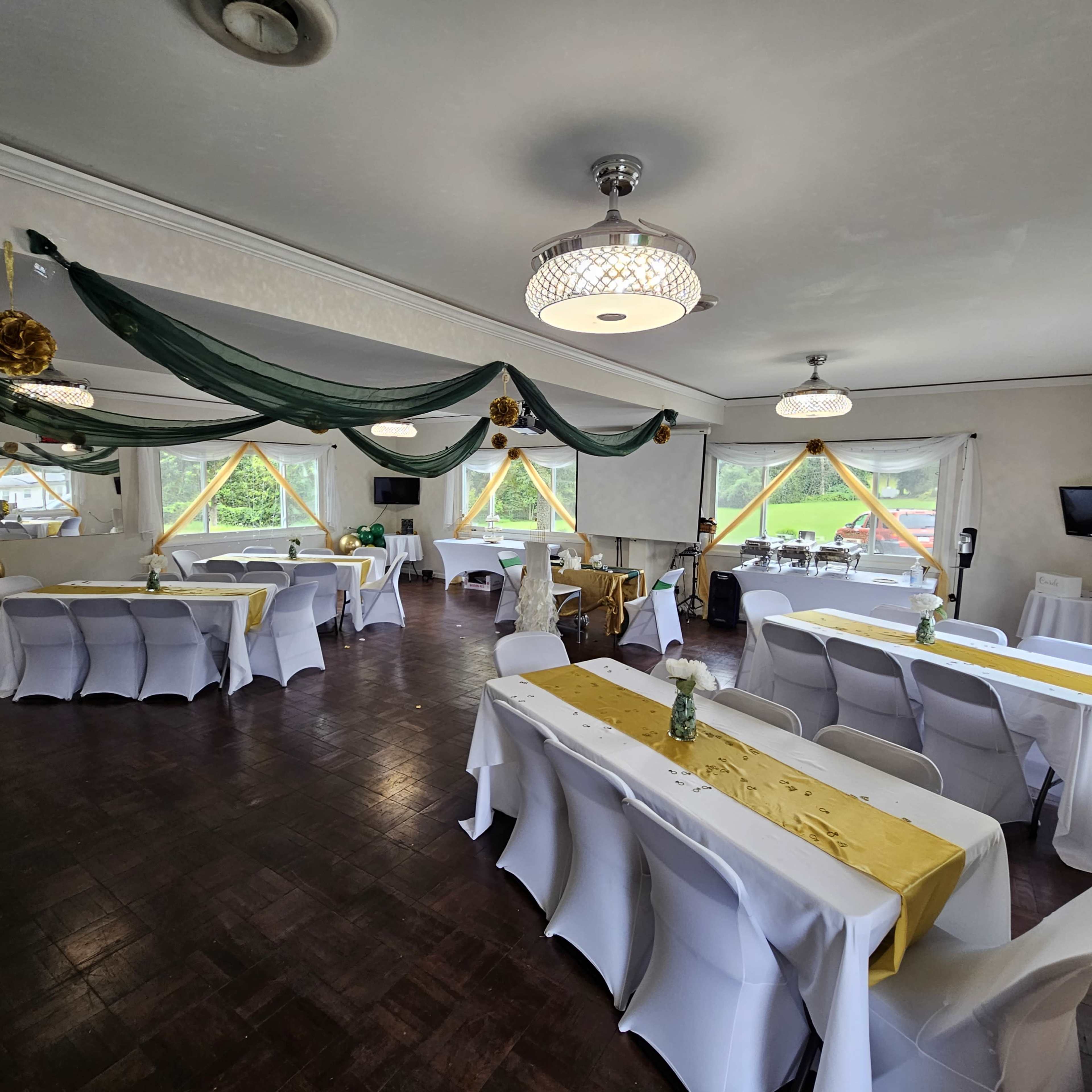 The image shows a banquet hall set up with white and yellow tablecloths, arranged with chairs, and decorated with drapes and ornaments.