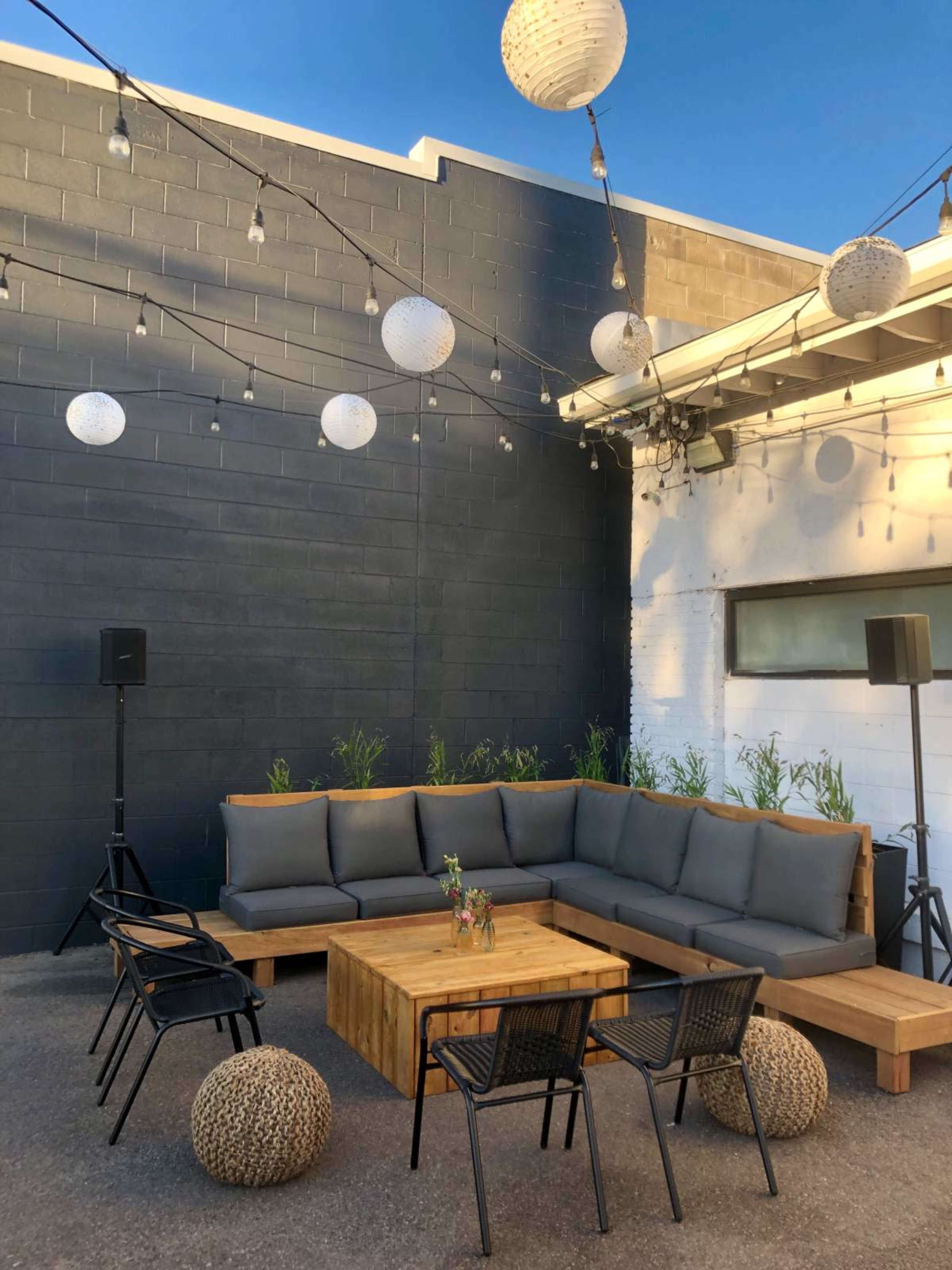 A cozy outdoor lounge area features a wooden sectional sofa, a low table, and black metal chairs, all arranged under decorative lanterns and string lights.