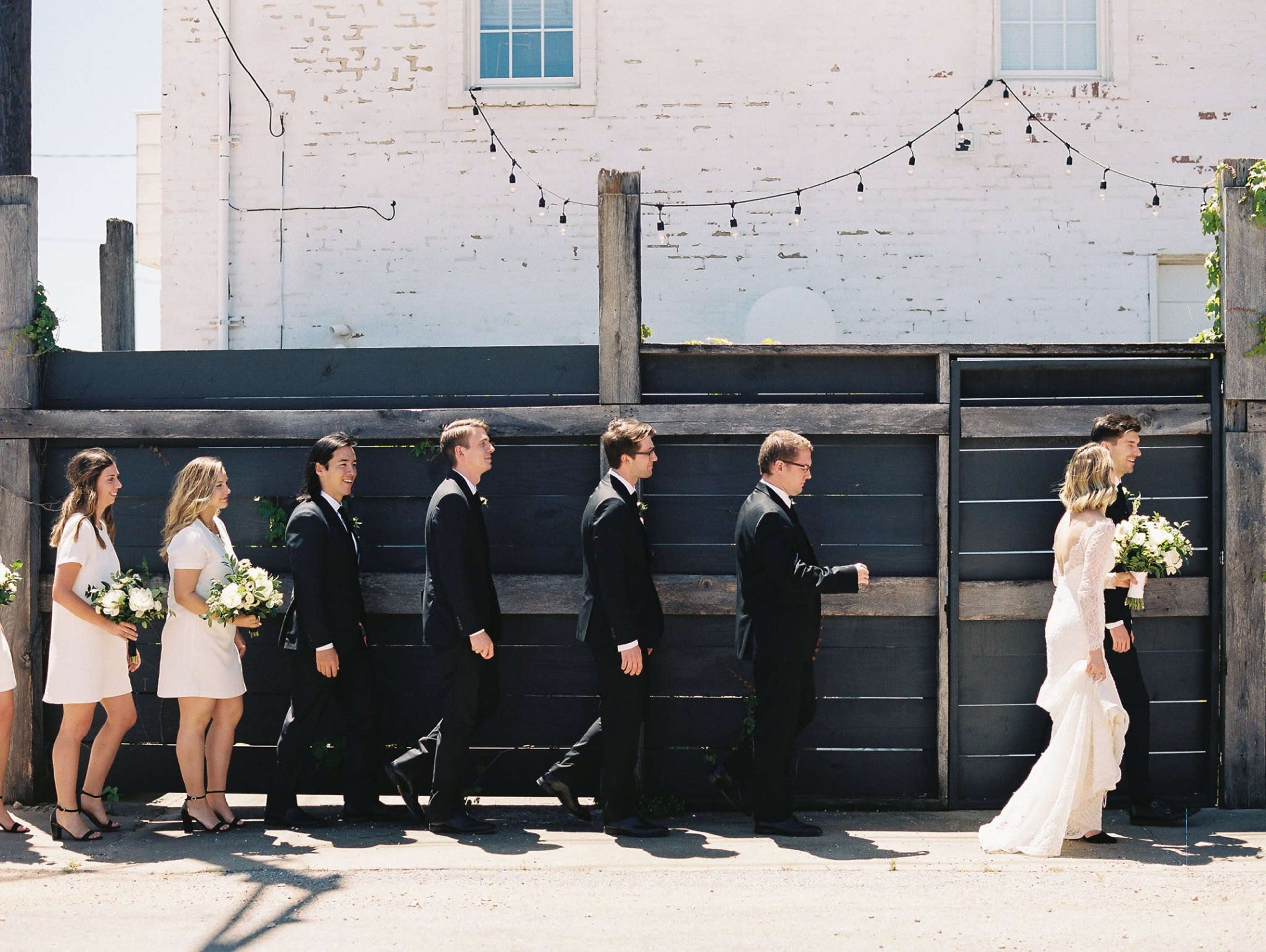 A wedding party, dressed in formal attire, walks in a line along a gray wooden fence.