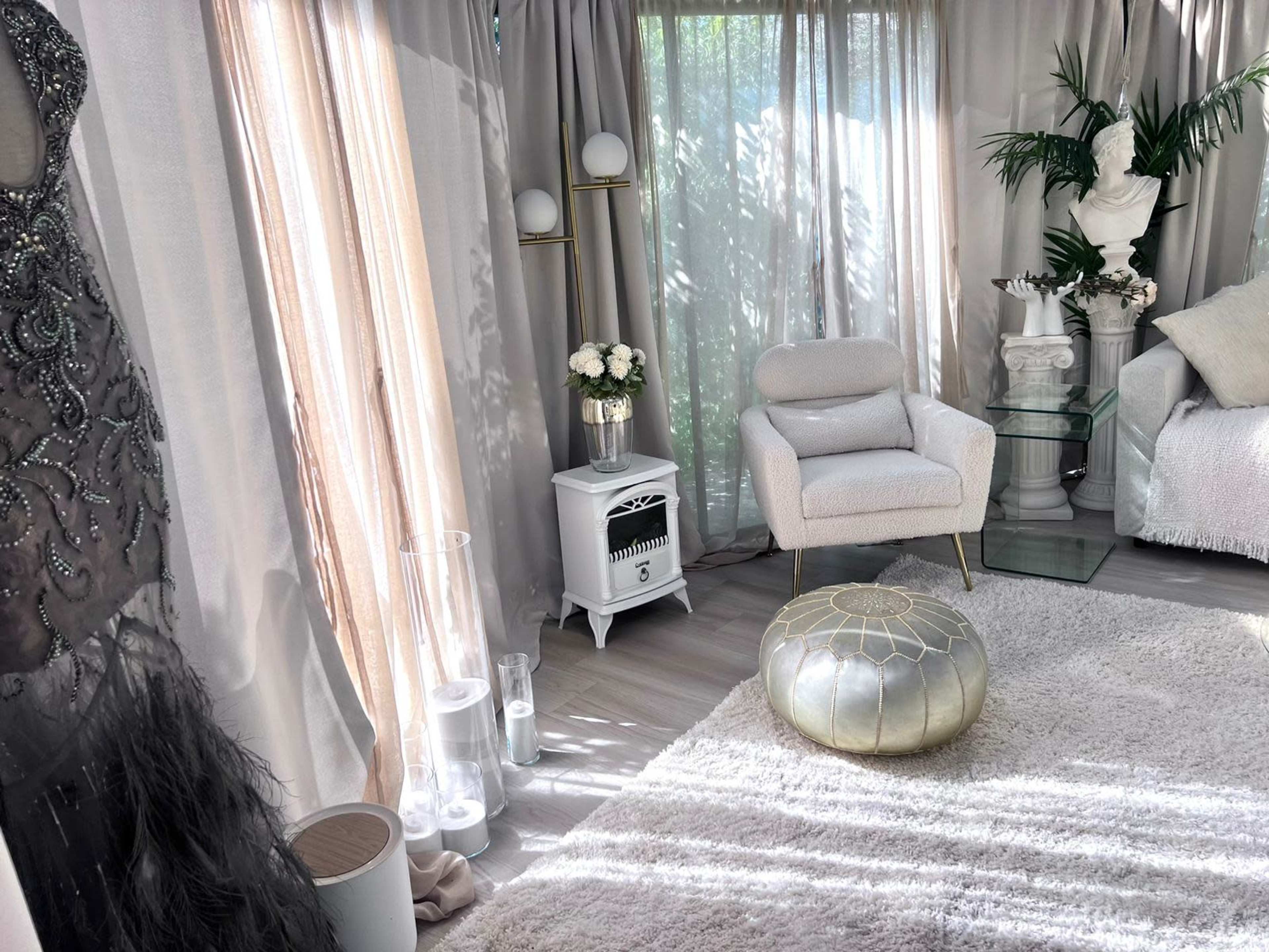 A stylish interior with a plush white rug, a glass side table, a white armchair, and a decorated corner featuring plants and soft lighting.