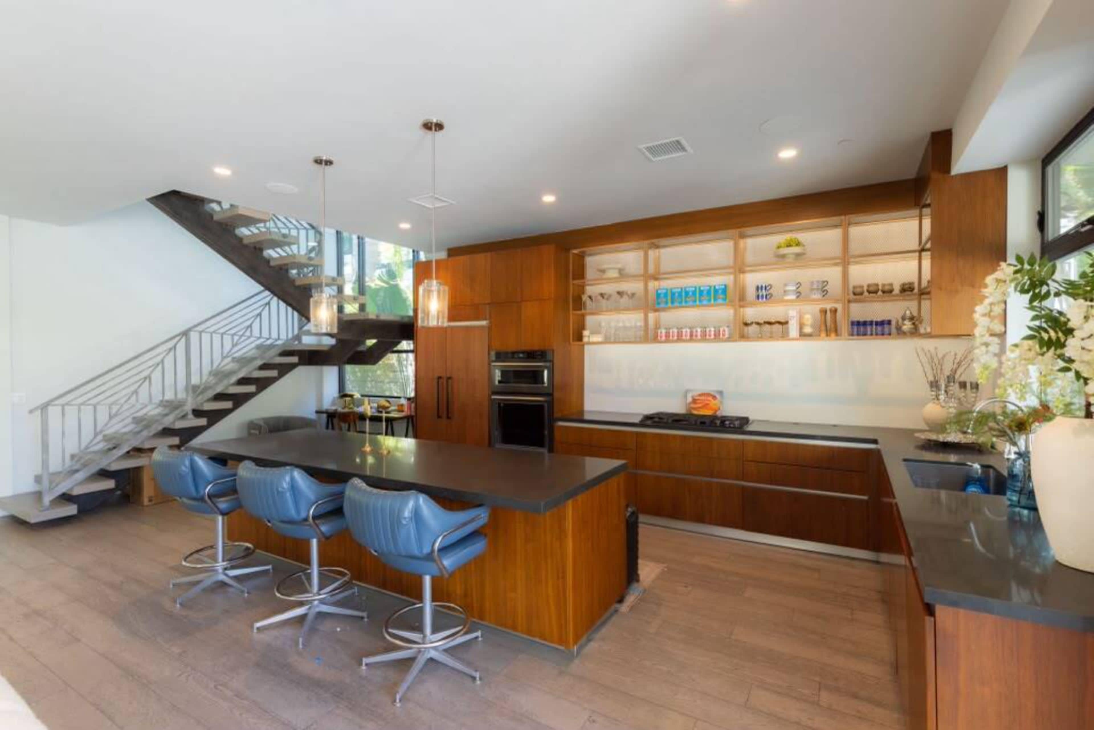 The image shows a modern kitchen featuring a large island with four seating stools, wooden cabinetry, and a staircase in the background.
