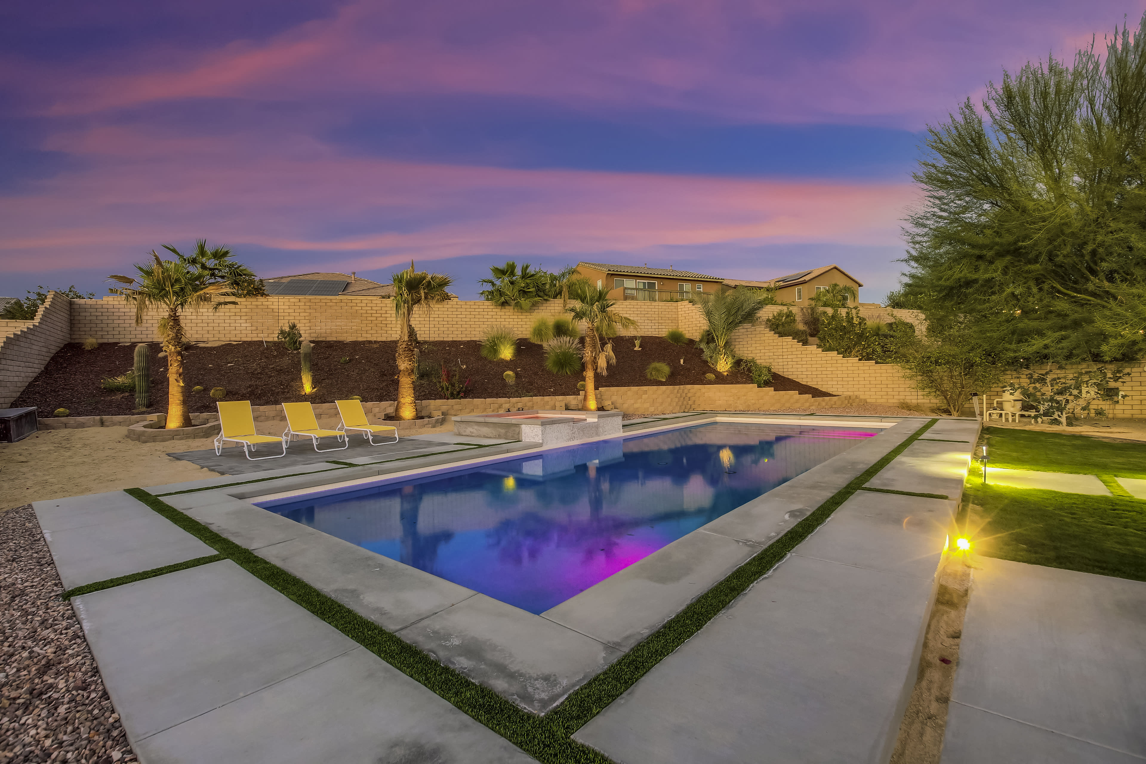 The image features a swimming pool with colorful lighting surrounded by palm trees and lounge chairs, set against a backdrop of a vibrant sunset sky.
