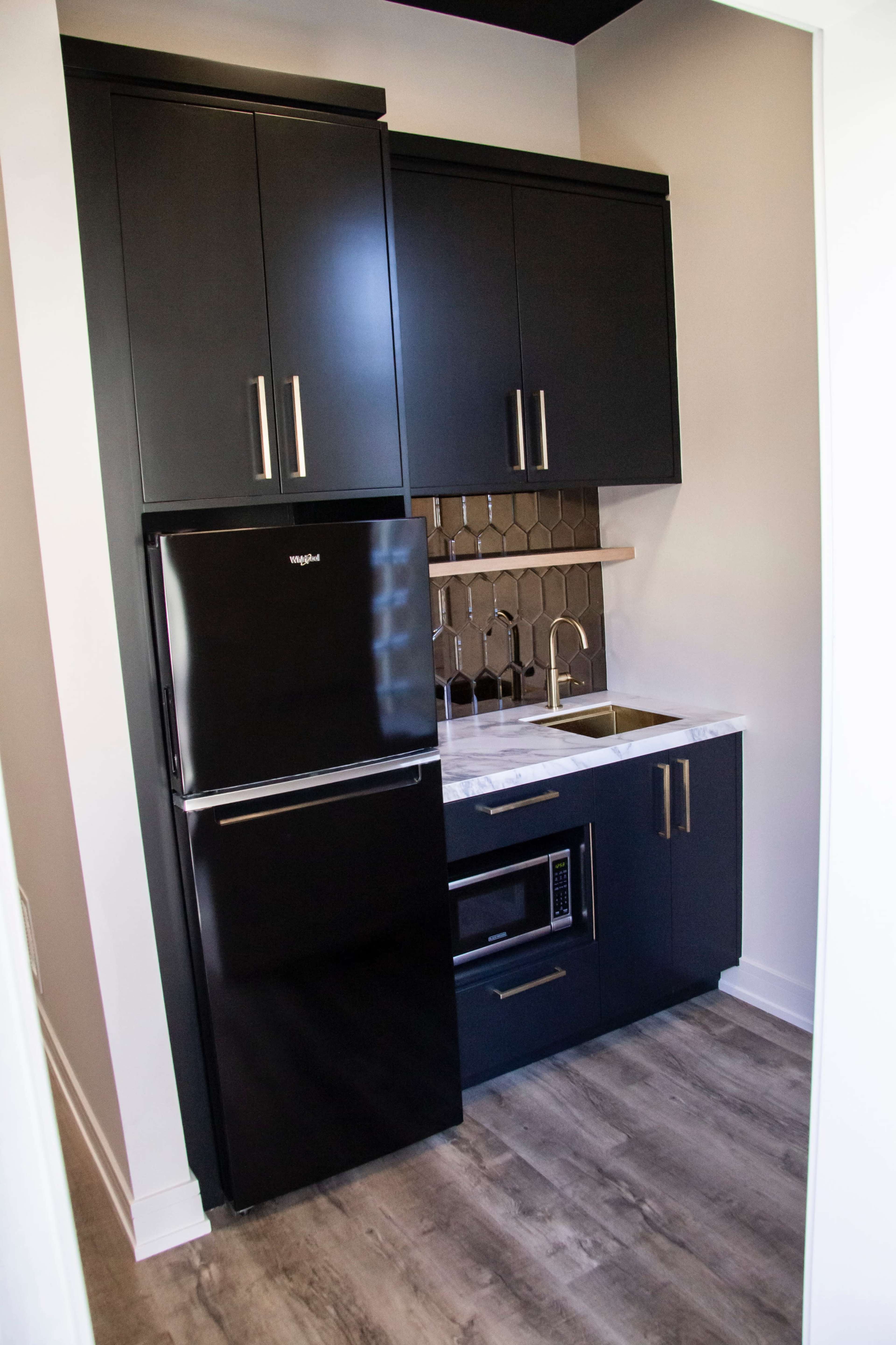 The image shows a compact kitchen area featuring dark cabinets, a black refrigerator, a sink, and a microwave beneath a hexagonal tile backsplash.