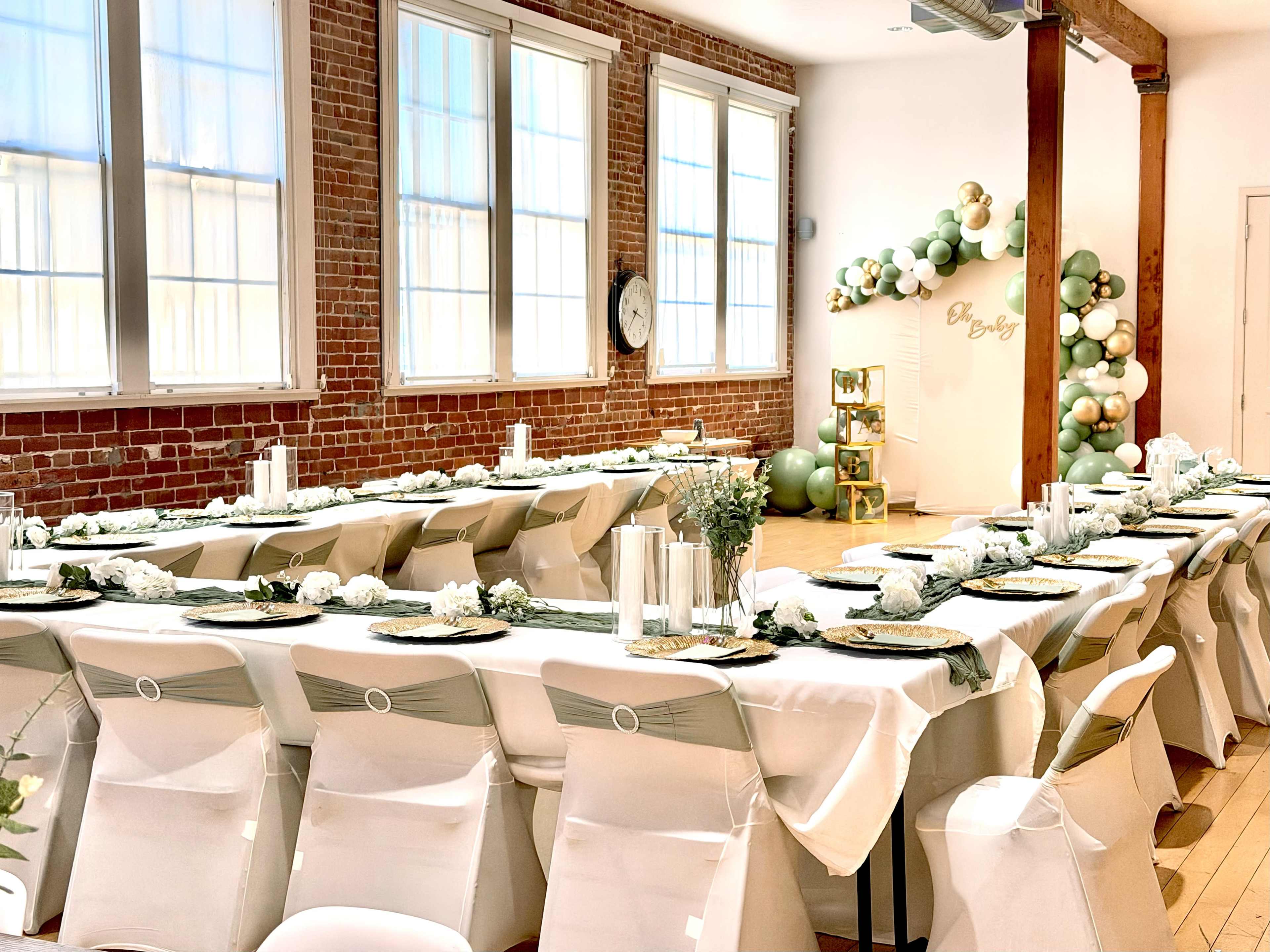 The image shows a decorated event space featuring long tables set with white tablecloths and green accents, alongside a wall adorned with balloons.