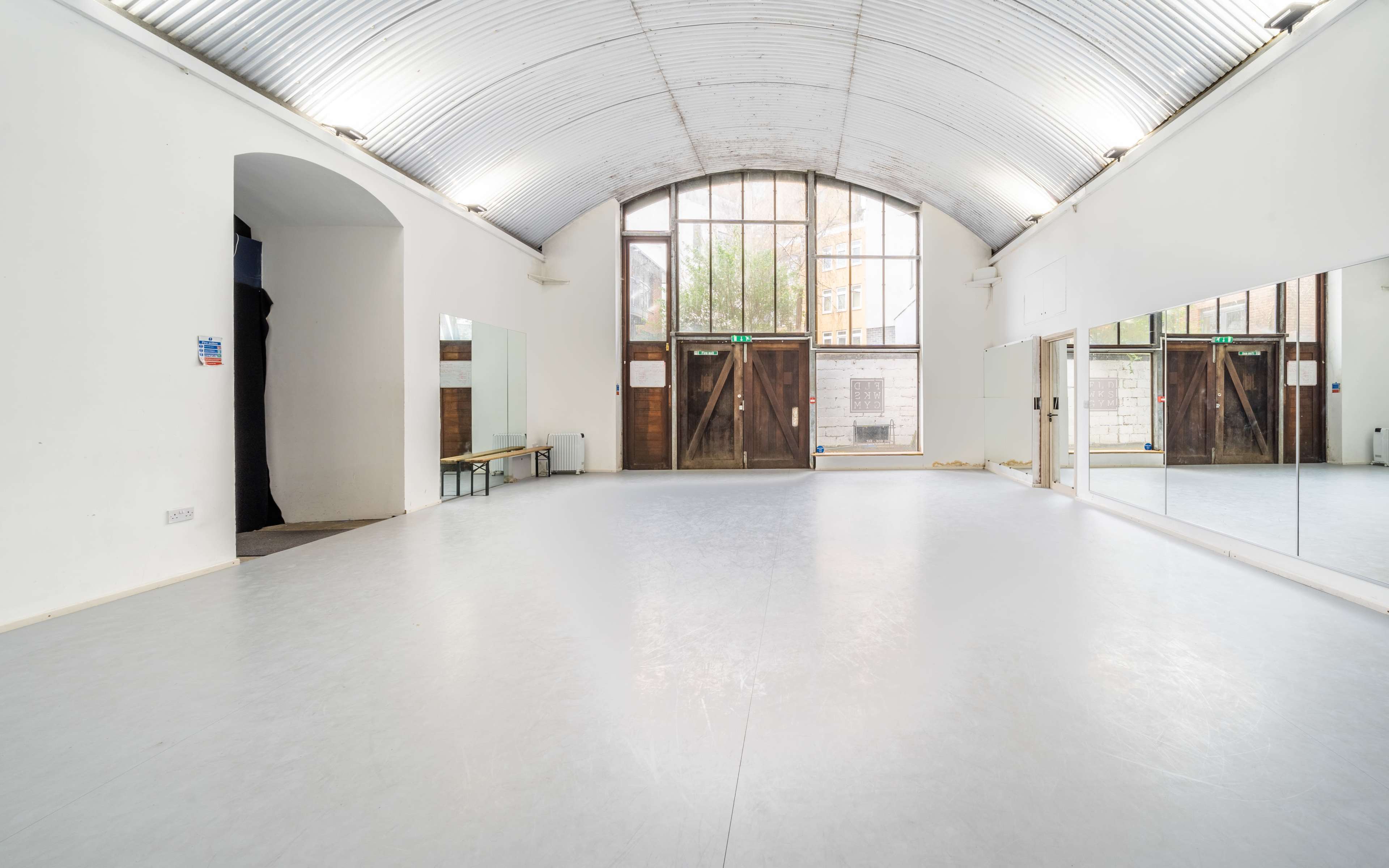 The image shows a spacious dance studio with a mirrored wall and large windows, featuring a light gray floor and wooden doors at one end.